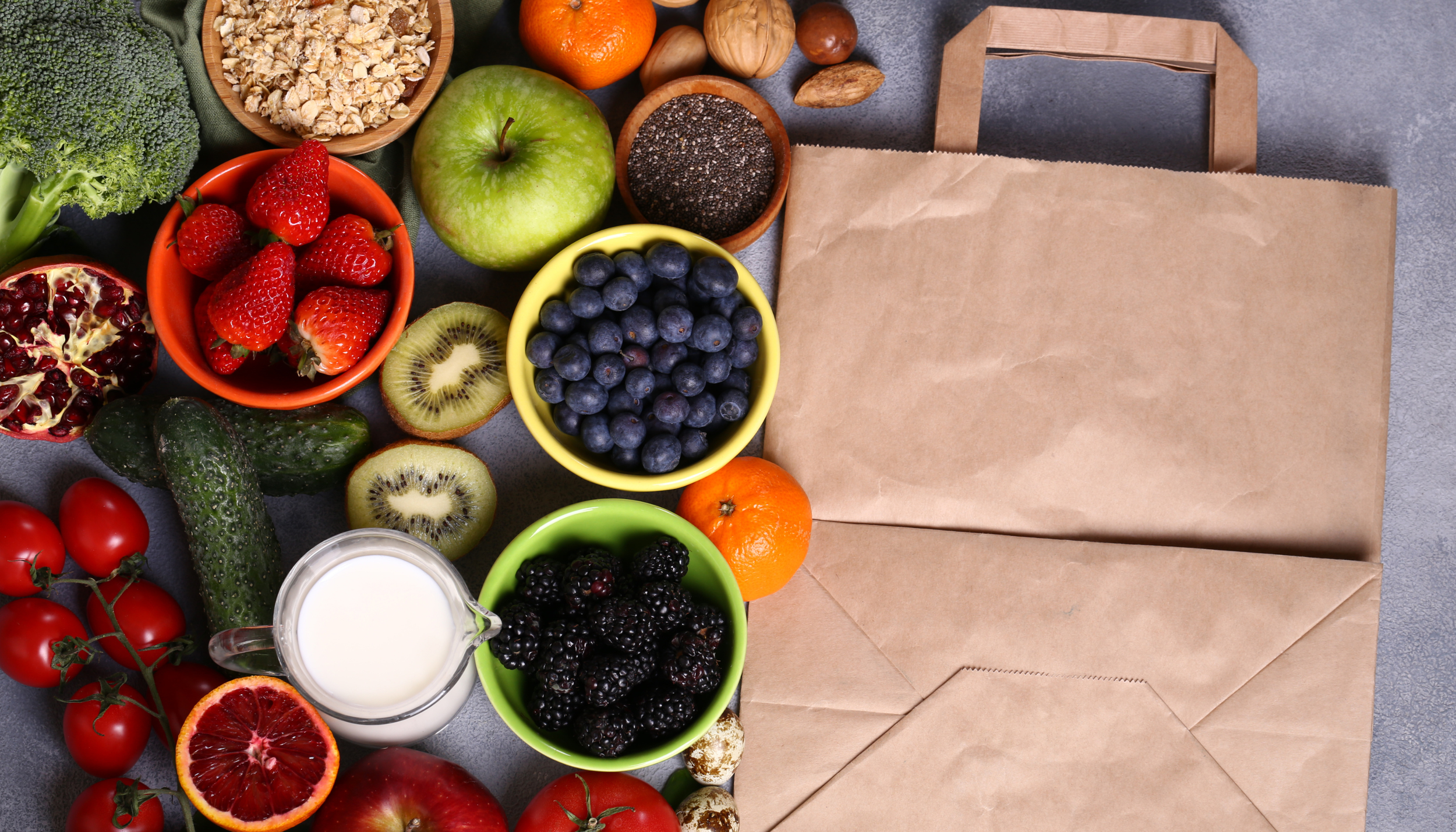A variety of fresh fruits including strawberries, blueberries, blackberries, grapes, kiwi, oranges, pomegranate, tomatoes, and cucumbers, along with bowls of chia seeds, oats, milk, and a slice of blood orange arranged on a table, with brown paper bags nearby.