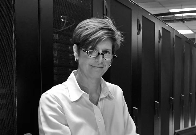 M/A Force Founder and CEO with short hair and glasses, wearing a white blouse, standing in a server room with black server racks.