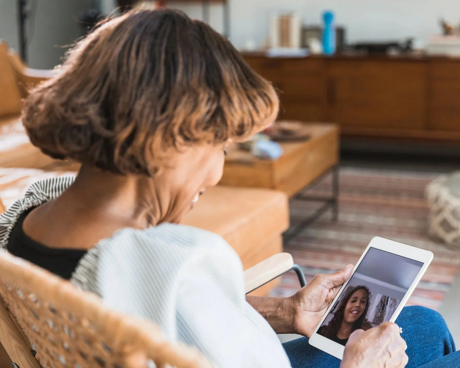 An elderly woman with short, light brown hair sitting on a wooden chair, holding a tablet and smiling at its screen during a video call.