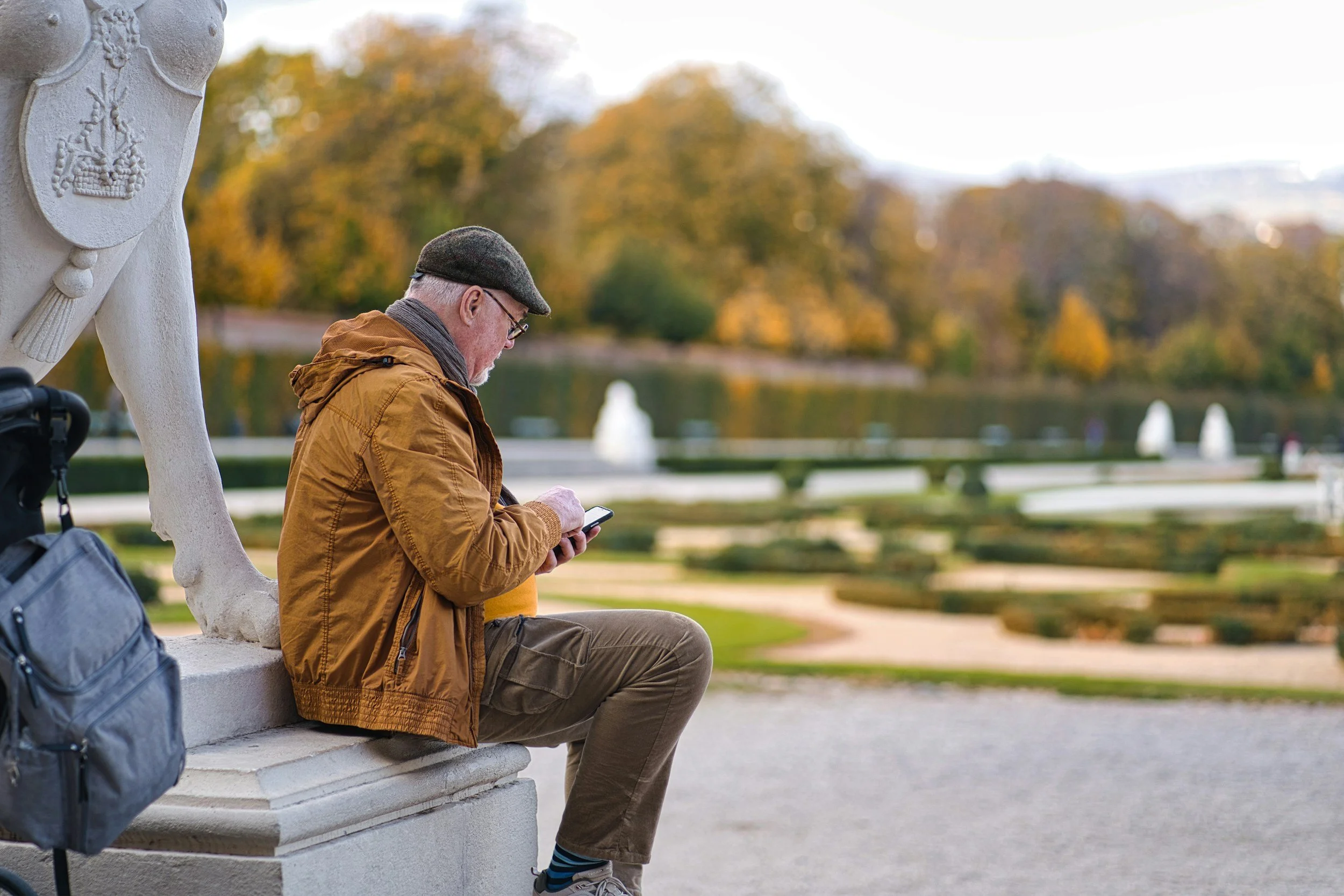 A man wearing a hat and tan coat uses a smartphone outside on a fall day.