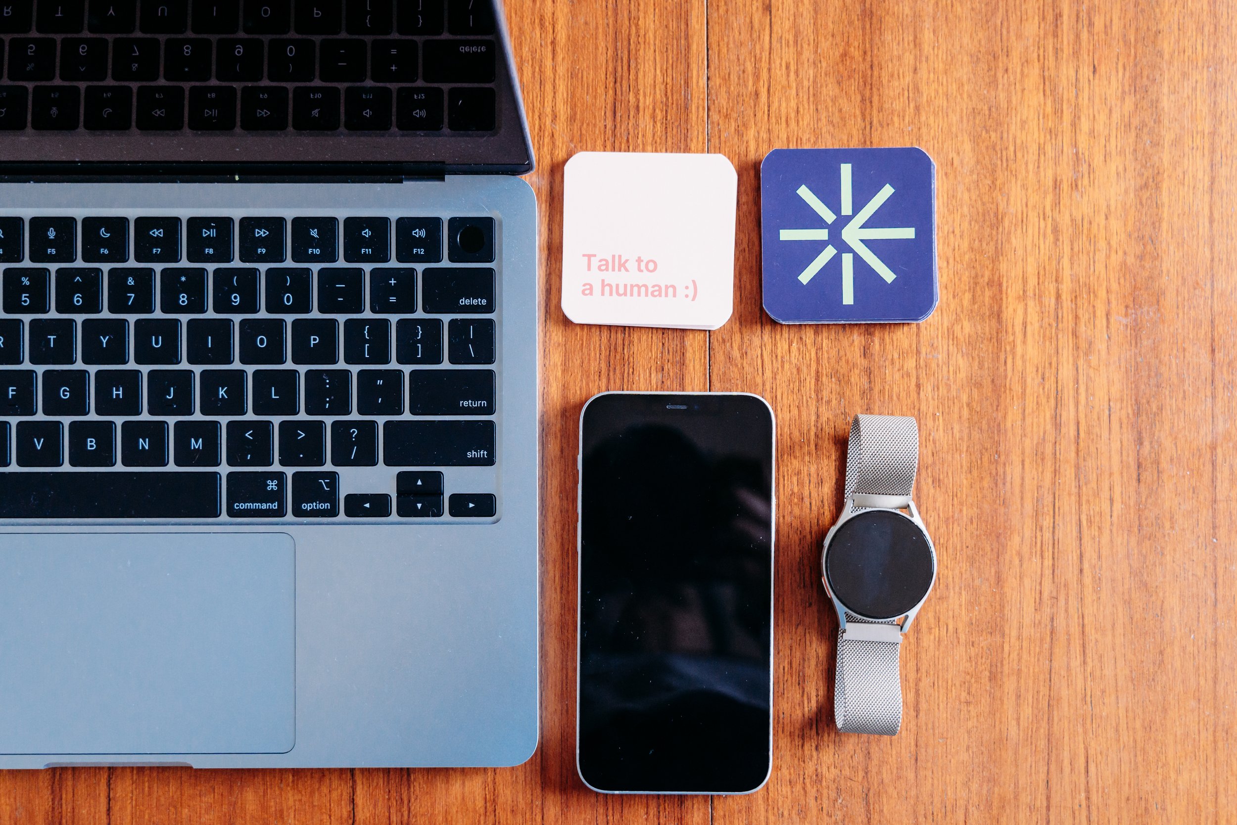 A wooden desk with a laptop, smartphone, smartwatch, two business cards, with one that says 'Talk to a human :)'.