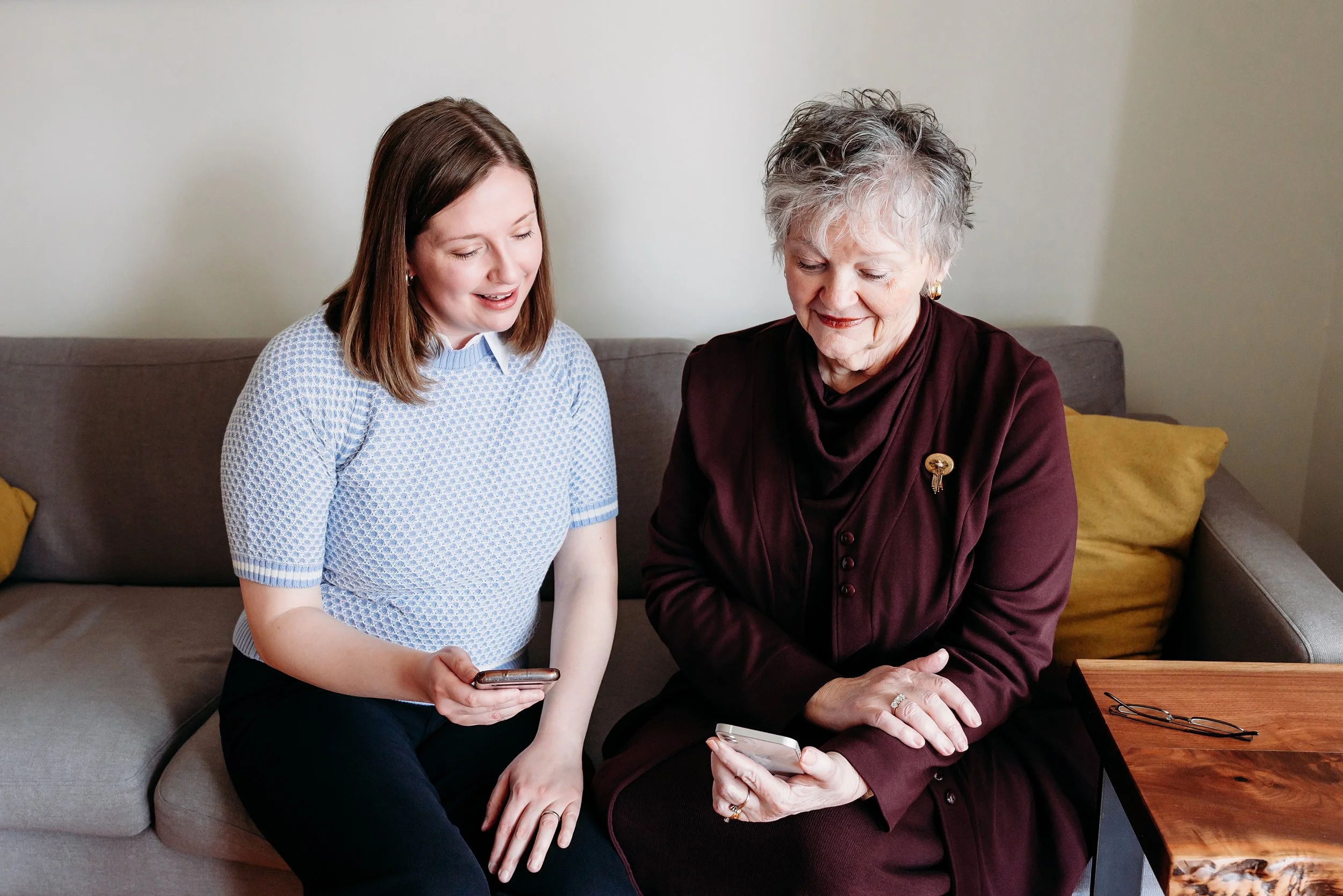 A young woman and an older woman sitting on a sofa, both looking at their smartphones and smiling.