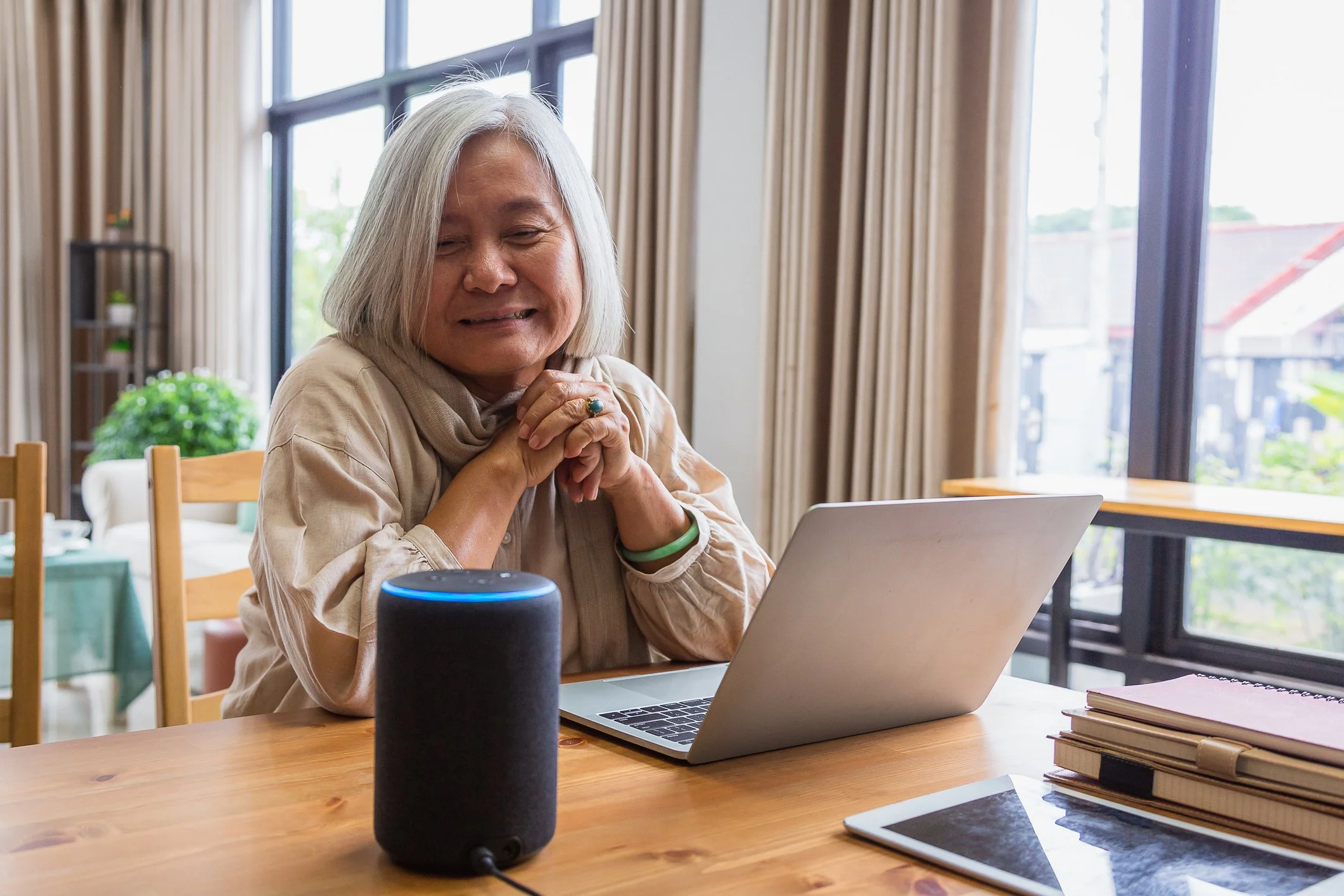 An elderly woman with white hair interacts with a smart speaker while sitting at a wooden table with a laptop, notebooks, and a tablet, in a room with large windows and natural light.