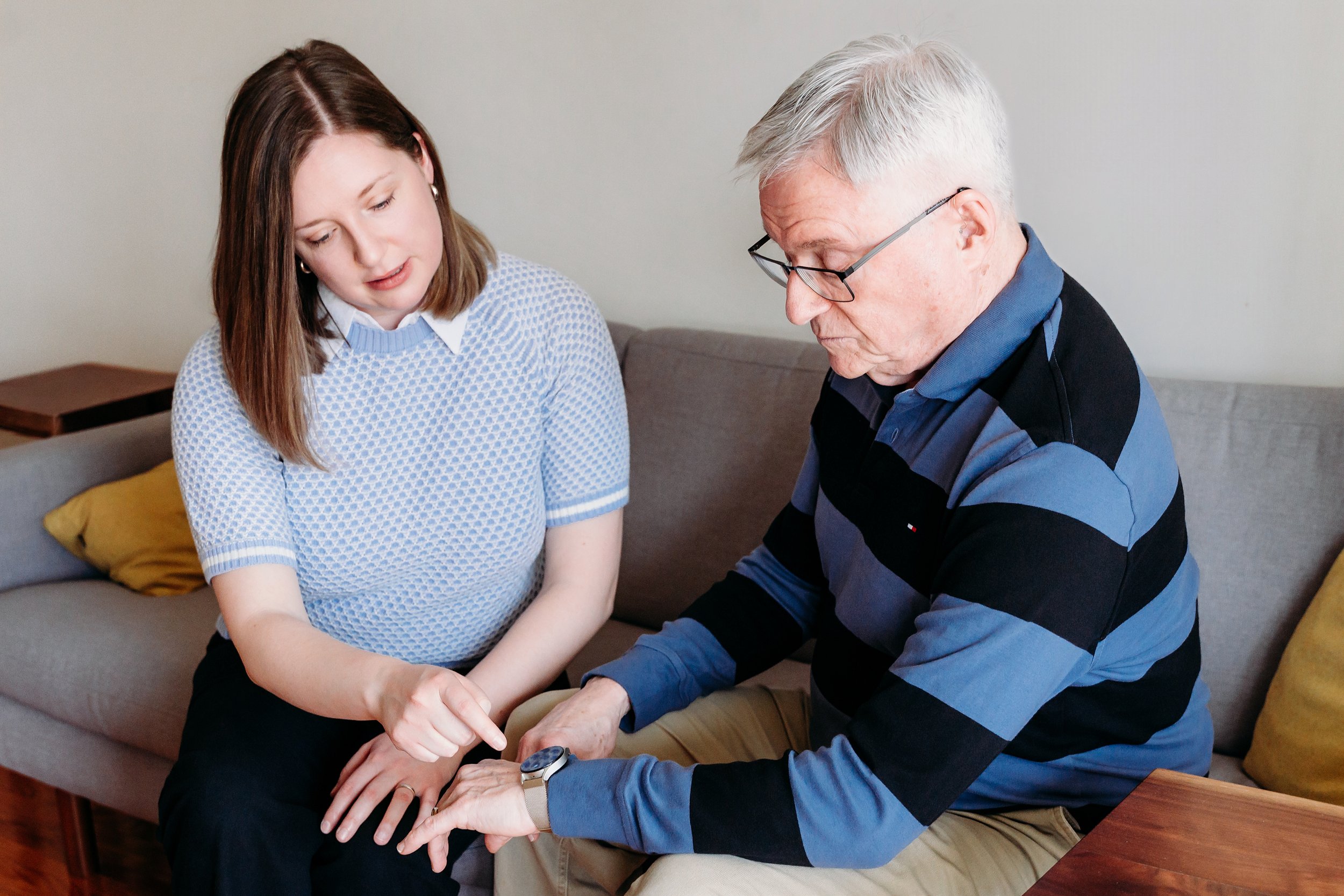 A woman showing an elderly man something on his smartwatch, while sitting on a sofa in a living room.
