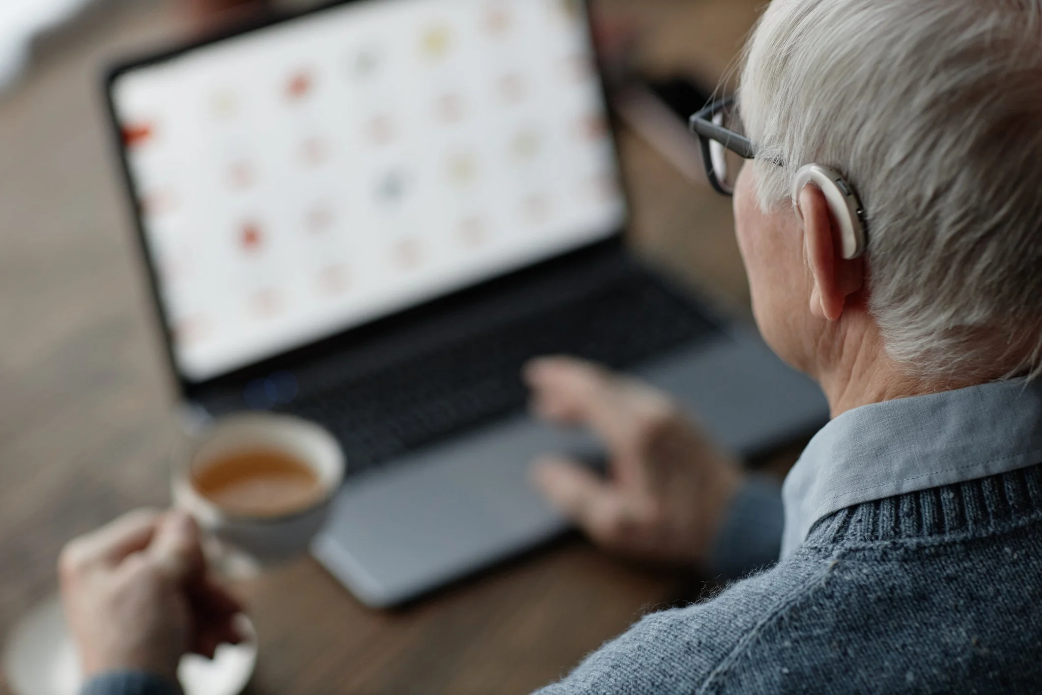 An elderly person with white hair and glasses wearing a hearing aid, sitting at a wooden desk, holding a cup of tea or coffee, and using a laptop computer.