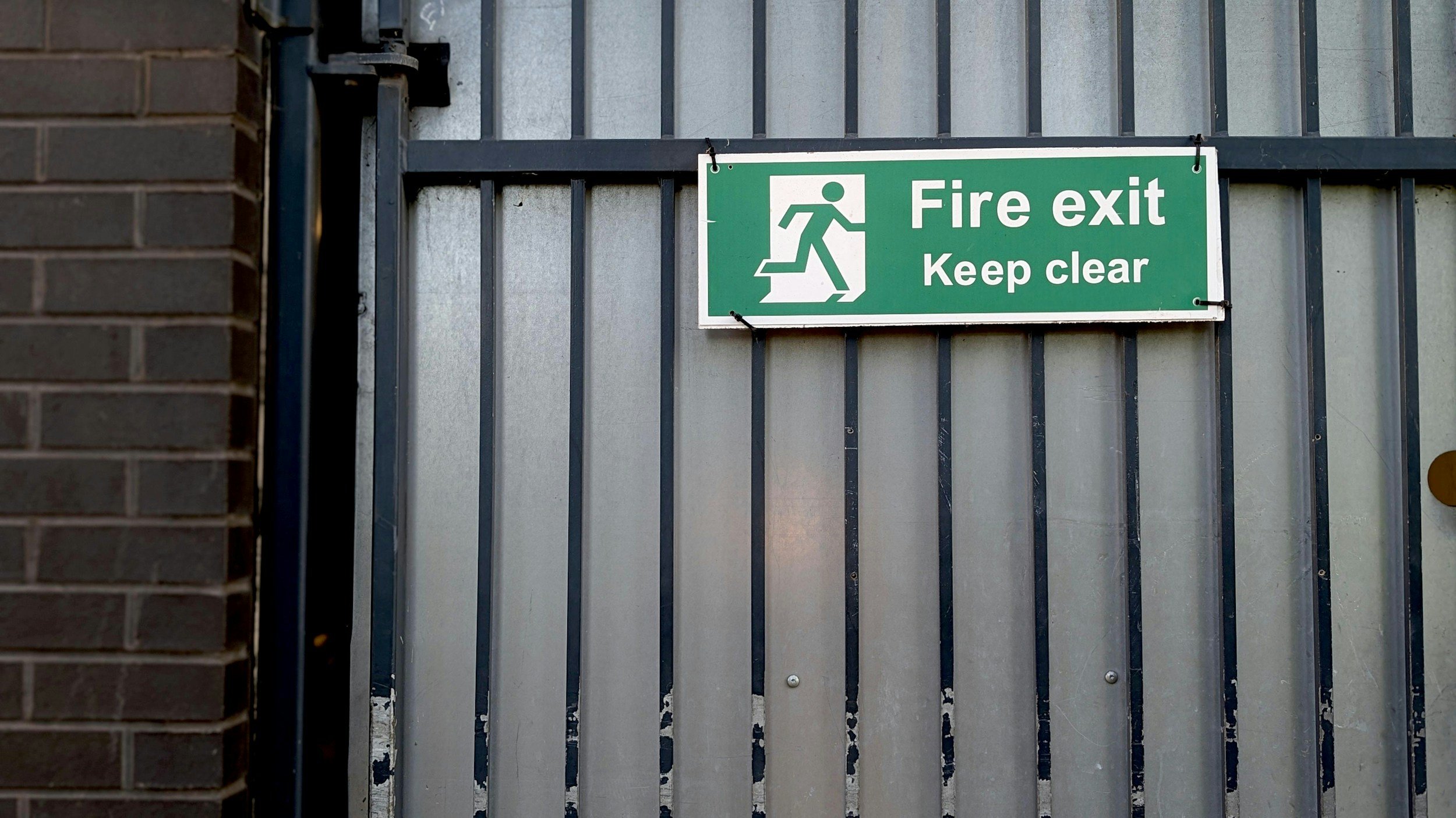 Green emergency exit sign with figure running and steps, mounted on a metal gate, reading 'Fire exit Keep clear'.