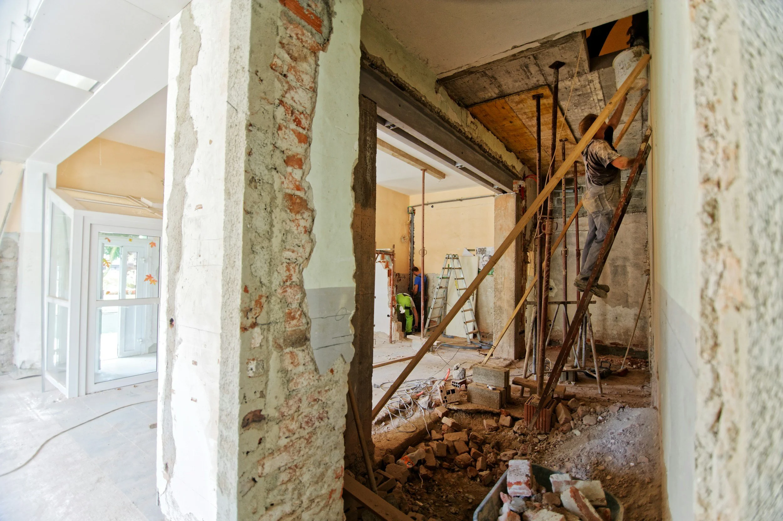 Construction workers renovating an interior space, with one worker on a wooden ladder and another worker in the background, surrounded by construction materials and debris.