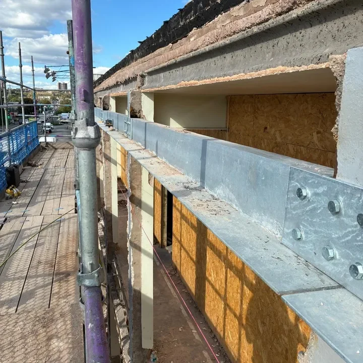 Construction site on a building with scaffolding, showing a metal rail or beam attached to the structure. Part of a walkway and street with vehicles are visible in the background.