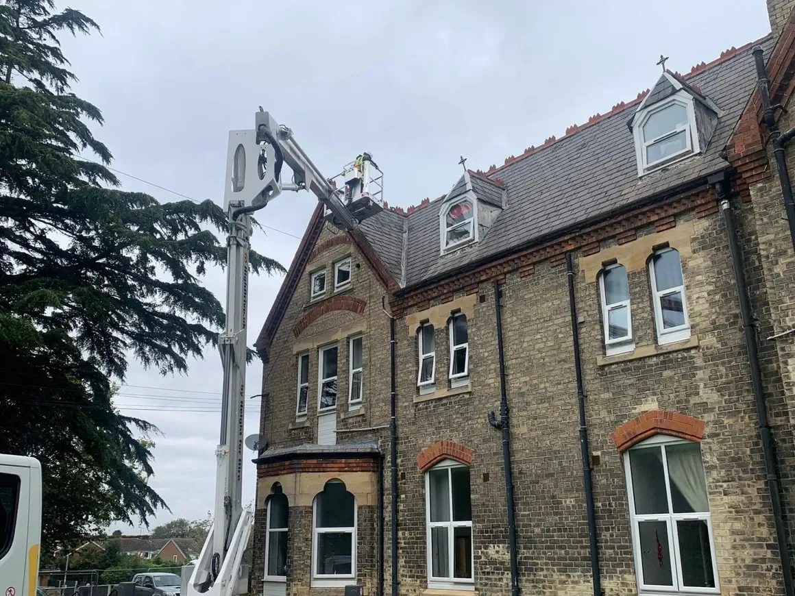 Workers on a cherry picker inspecting or repairing the roof of a brick house with dormer windows.