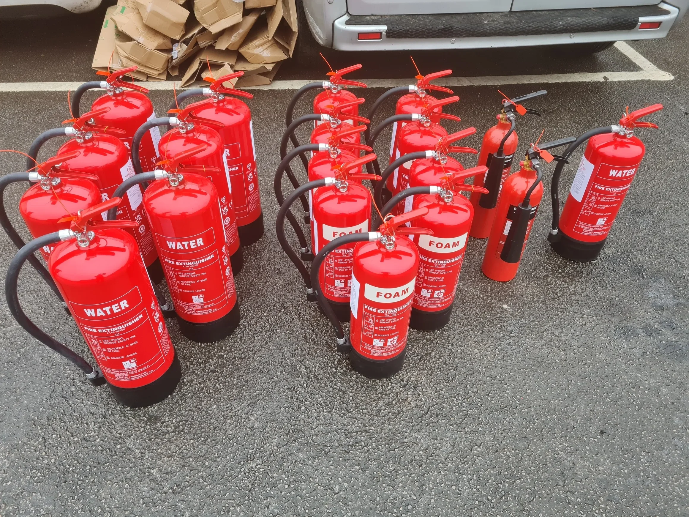 Multiple fire extinguishers labeled for water and foam, arranged on an outdoor asphalt surface near a parking space, with a silver vehicle and cardboard boxes in the background.