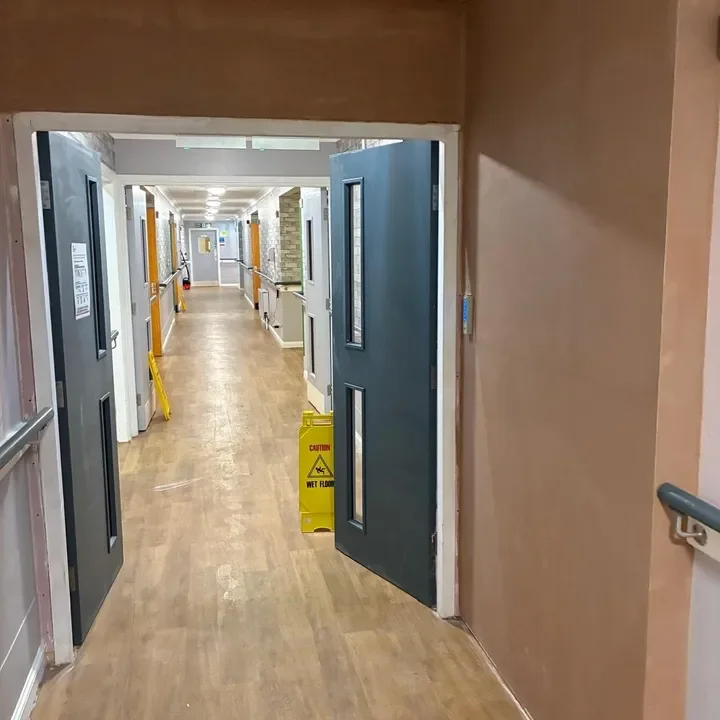 Hospital corridor with open door, yellow caution signs on wet floor, and hallway extending into the distance.