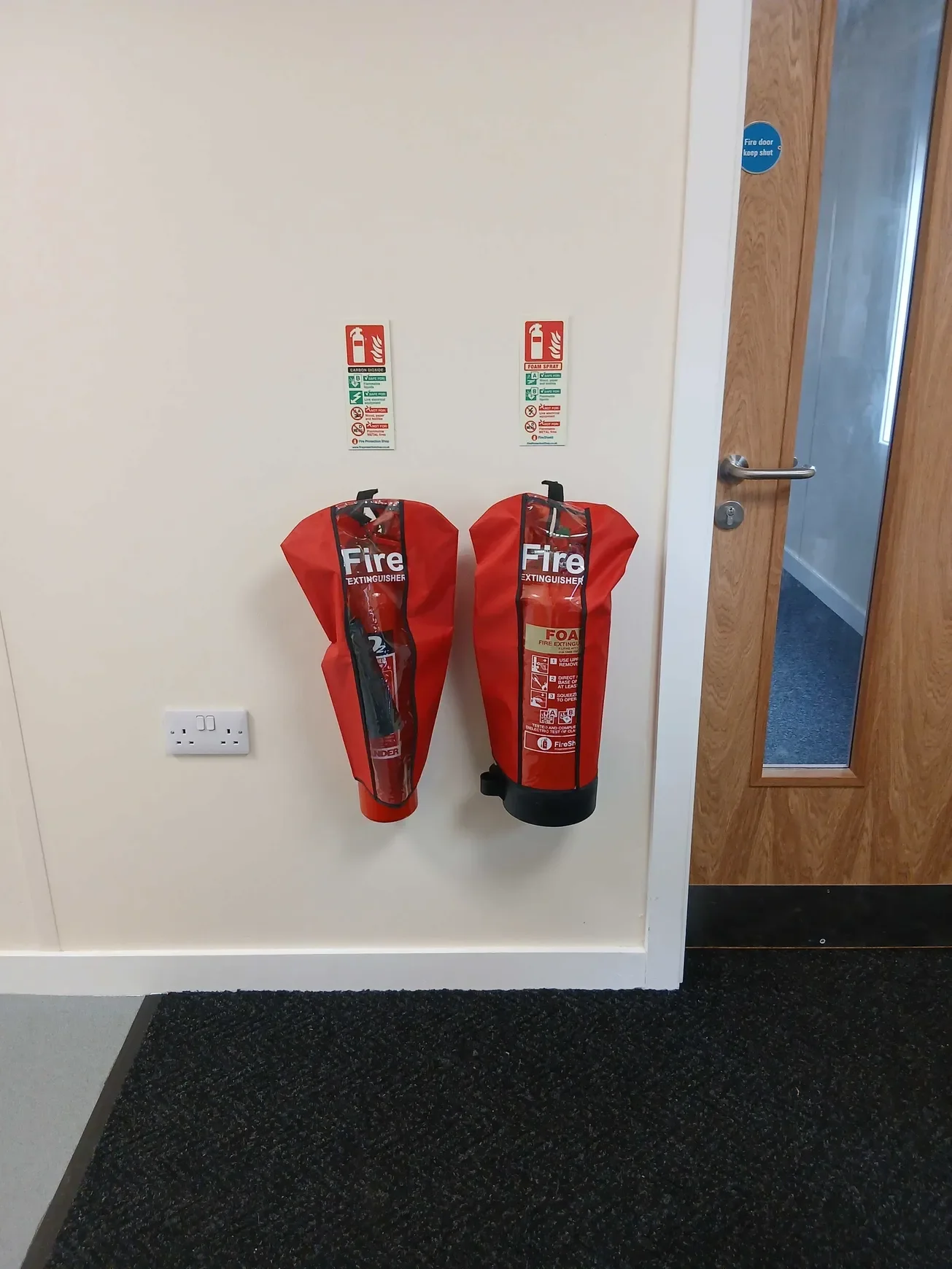 Two red fire extinguisher bags with instructions are mounted on a beige wall next to a wooden door with a glass panel.