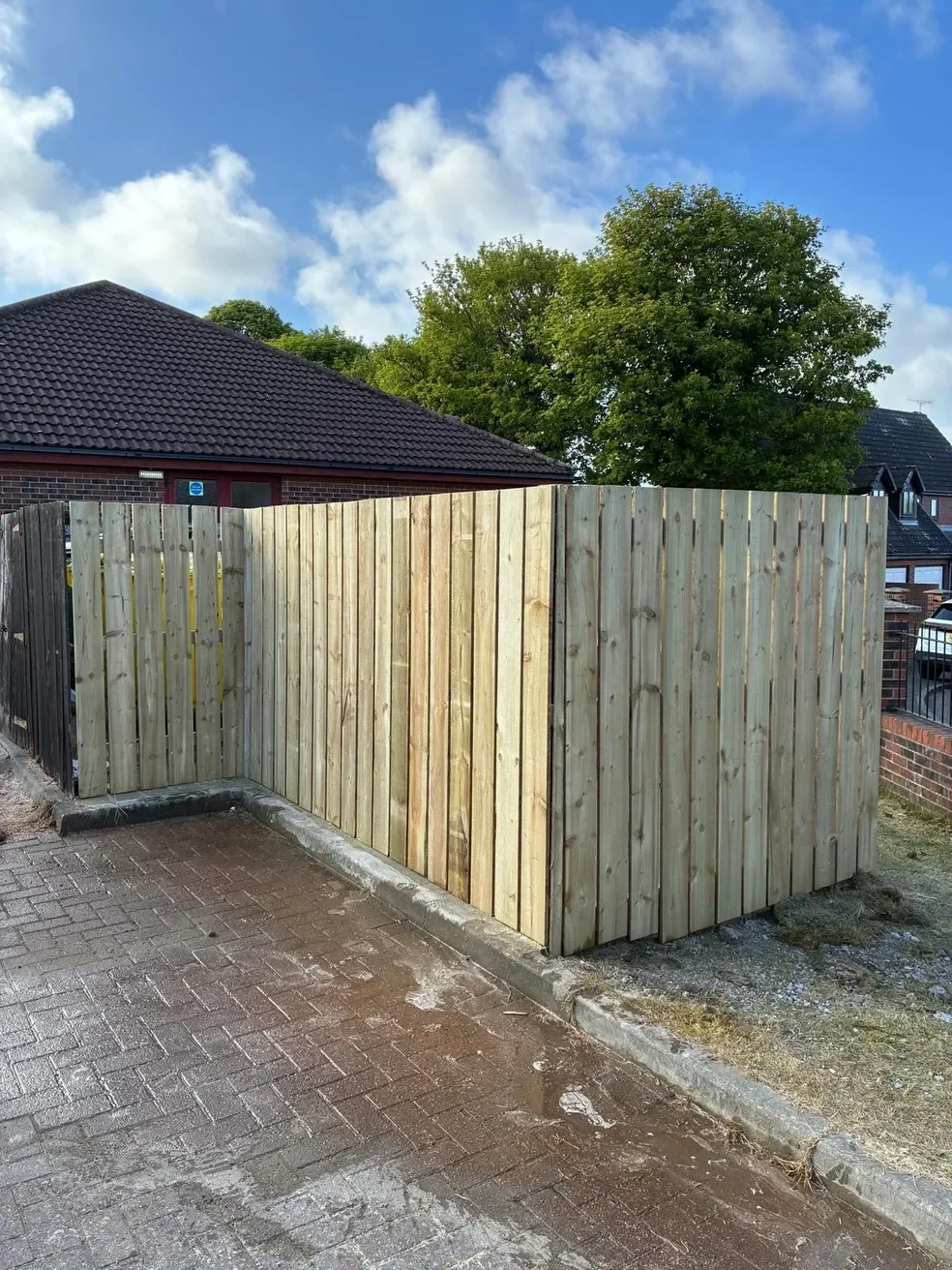 newly installed wooden privacy fence with different shades of wood panels surrounding a corner on a paved surface with a house and tree in the background.