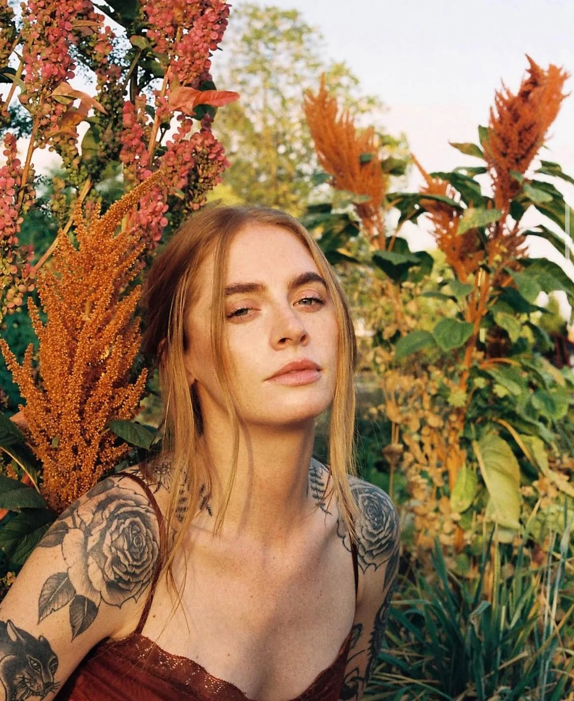A young woman with long hair and tattoos on her arms poses outdoors among colorful autumn plants and flowers.