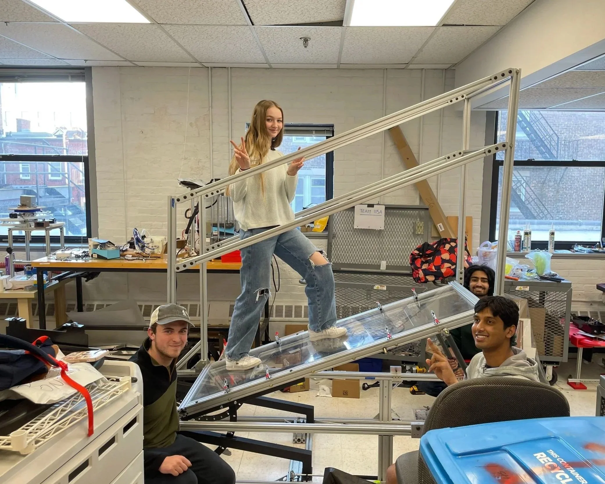 A young woman standing on a ramp structure inside a workshop, posing with peace signs. Four young men are around her, with some smiling and pointing at the camera. The workspace has windows, tools, and various objects, indicating a tech or engineerin