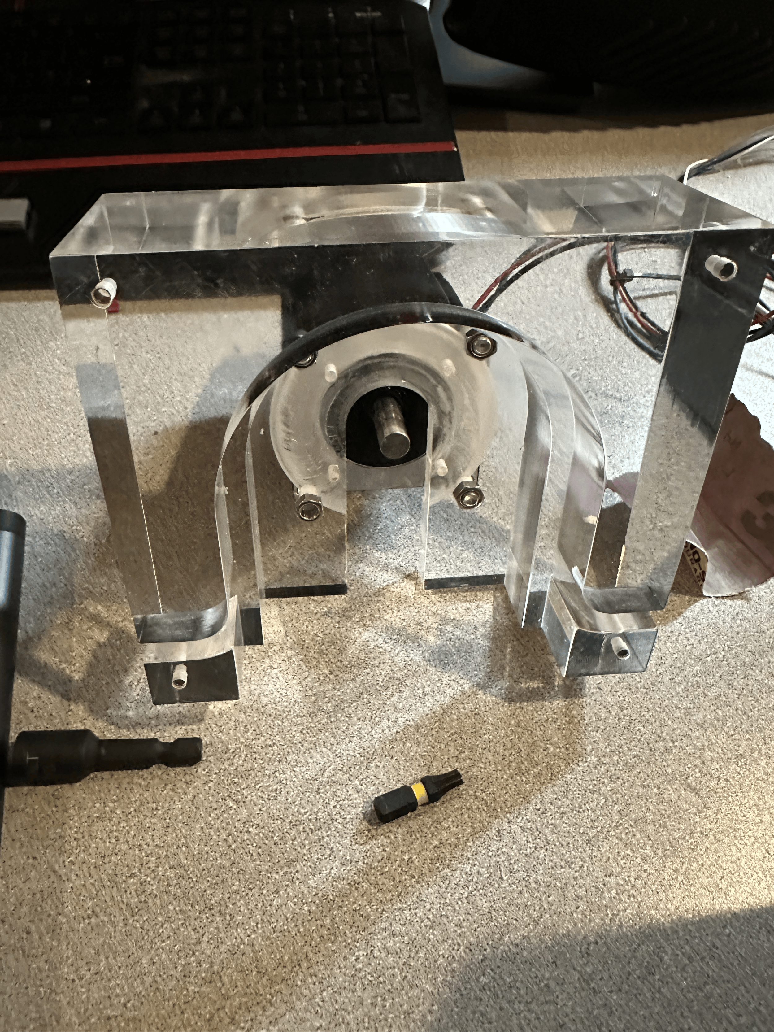 A clear acrylic motor mount with a black shaft, attached to a metal frame, placed on a beige carpeted surface with a screwdriver nearby and a black keyboard in the background.