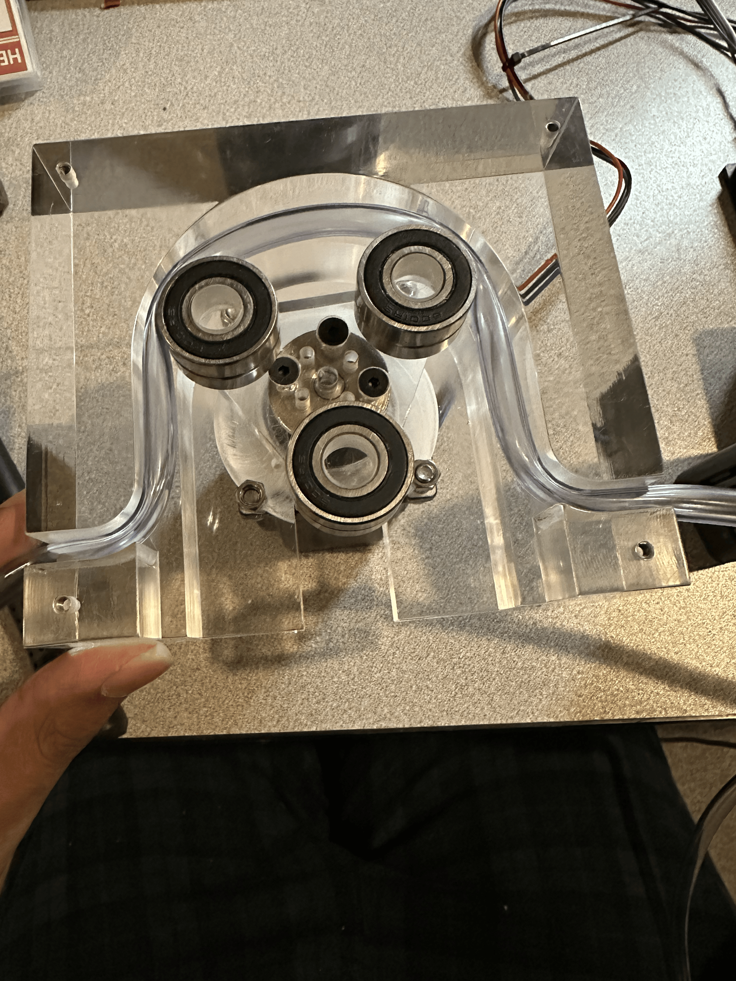 A transparent acrylic mechanical device with three visible bearings mounted on a metal frame, placed on a gray textured surface.