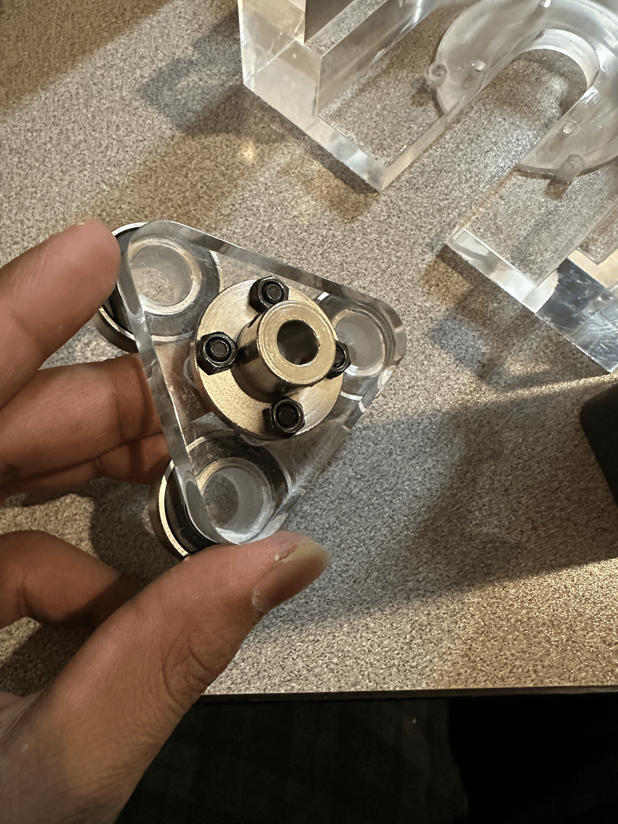Person holding a metal mechanical part with gears and bolts on a textured gray surface, surrounded by clear acrylic or glass blocks.