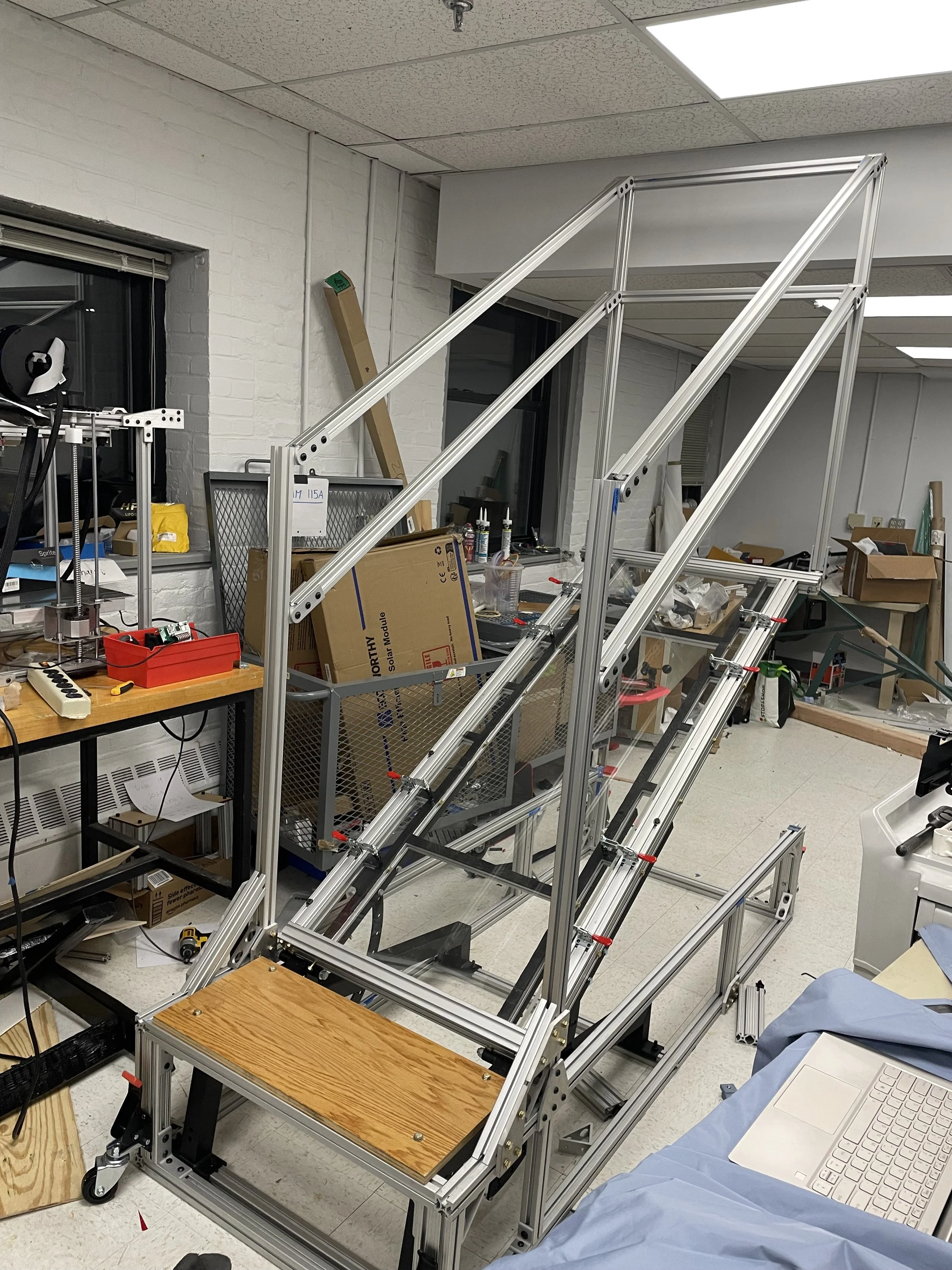 A metallic robot frame on wheels with multiple ramps and a wooden platform in a workshop or laboratory setting.