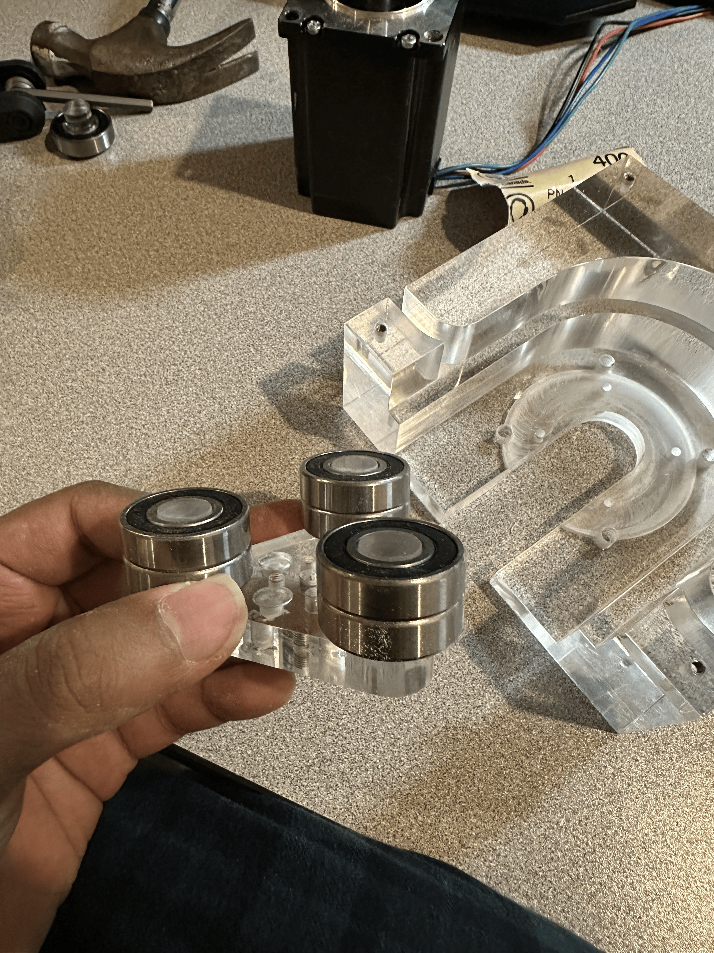 A person's hand holding a clear acrylic part with three small ball bearings on top of it, on a workbench with various mechanical parts and tools.