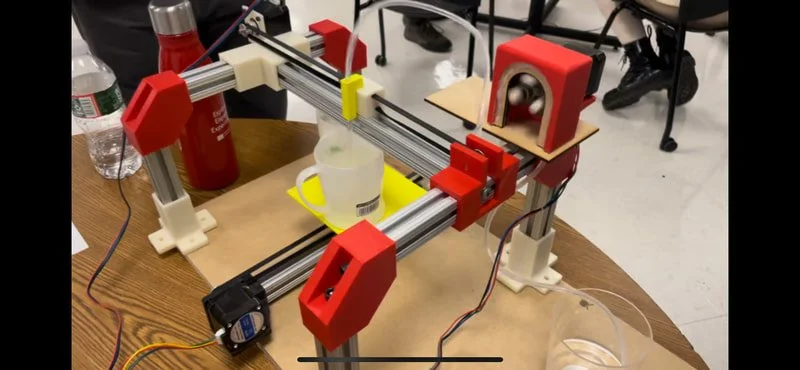 A 3D printer or CNC machine with red and white frame components, metal rails, and an extruder or cutting tool, set up on a wooden table with a partially printed or cut object.