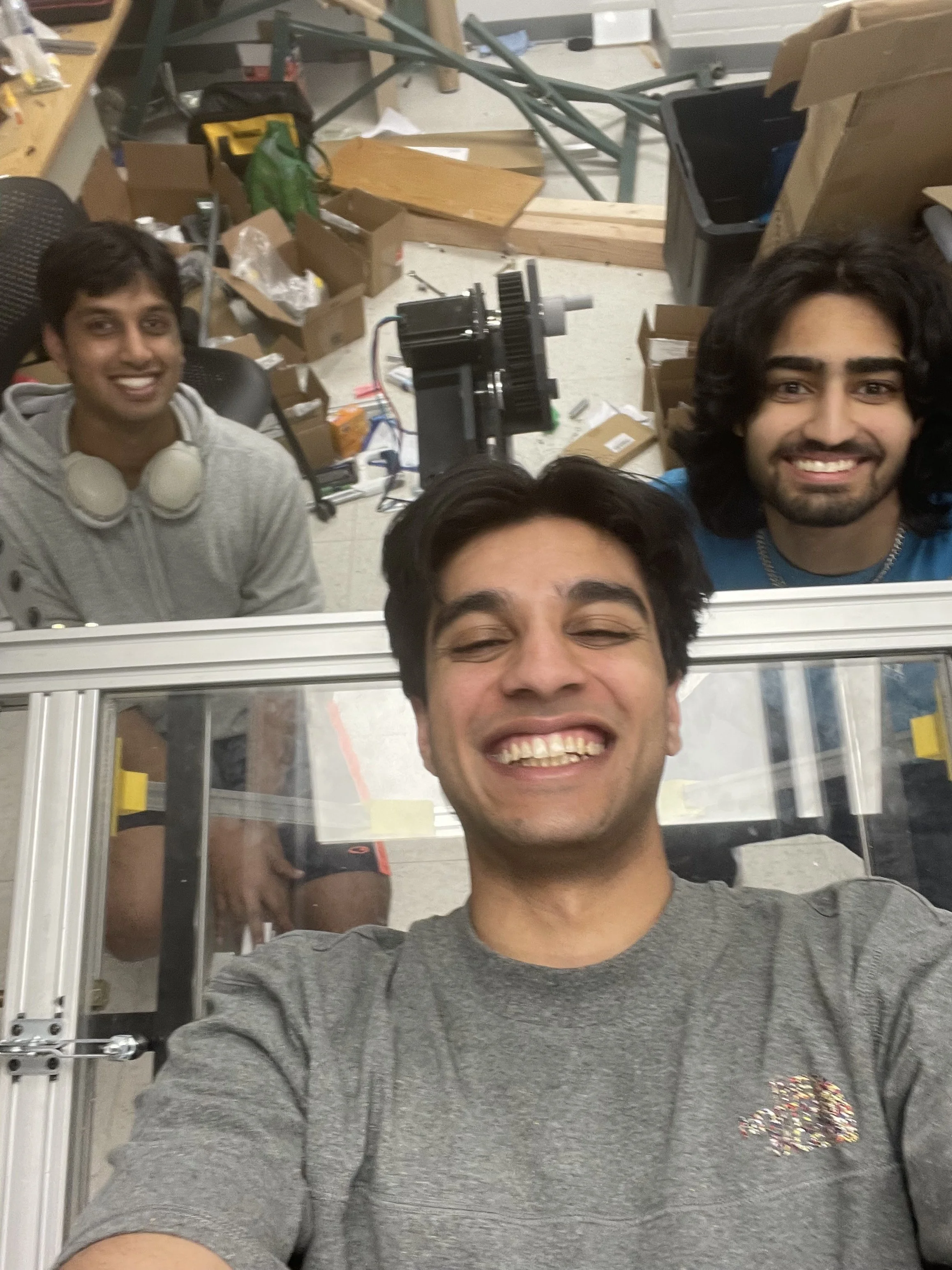Three smiling young men taking a selfie in a workshop or laboratory with various equipment, boxes, and tools in the background.
