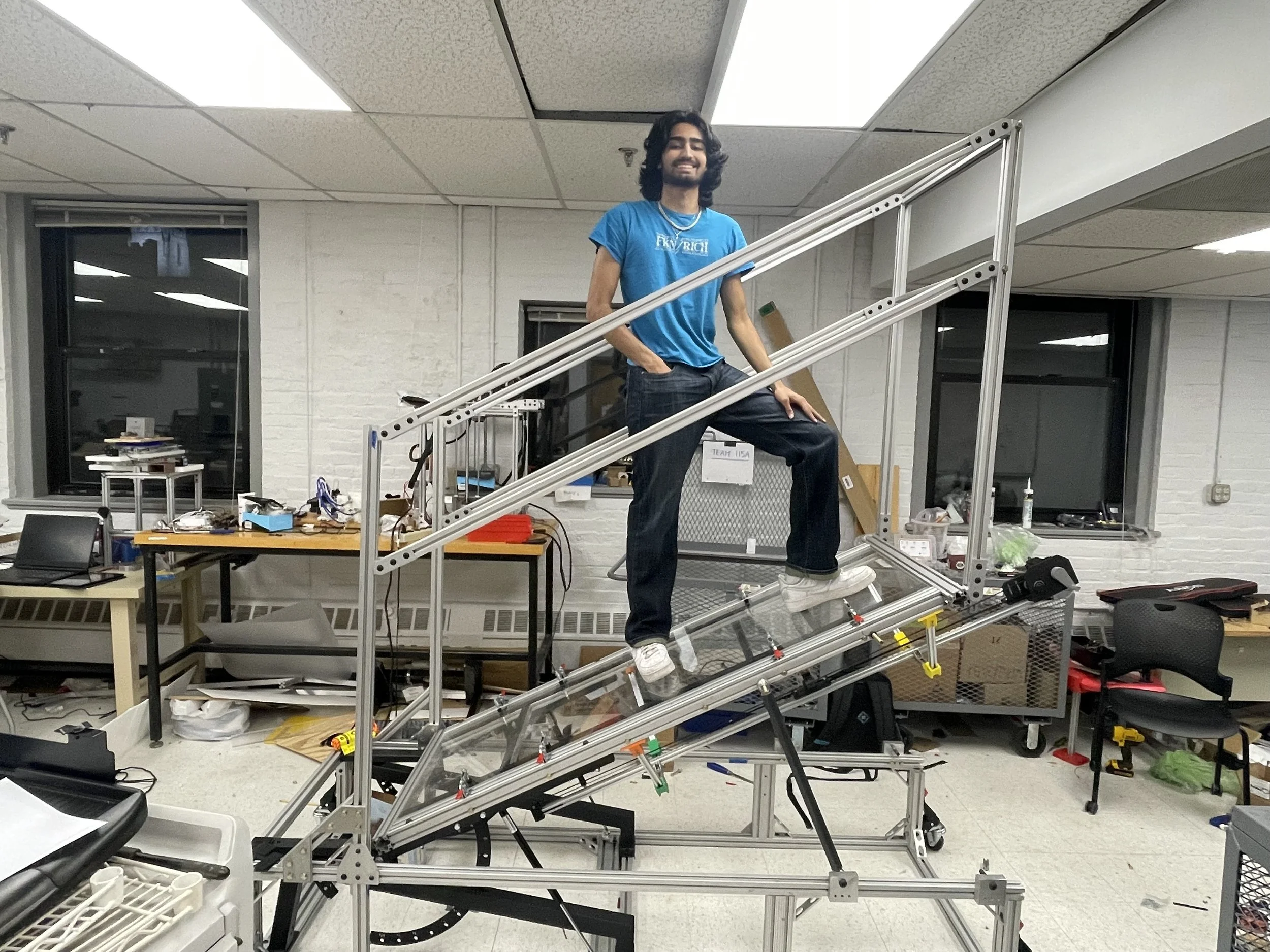 A person with long dark hair and a beard stands on a robot arm with a mechanical frame in an office or workshop. The person is smiling and wearing a blue T-shirt and jeans, with one hand on their hip and the other resting on the robot. The room has w