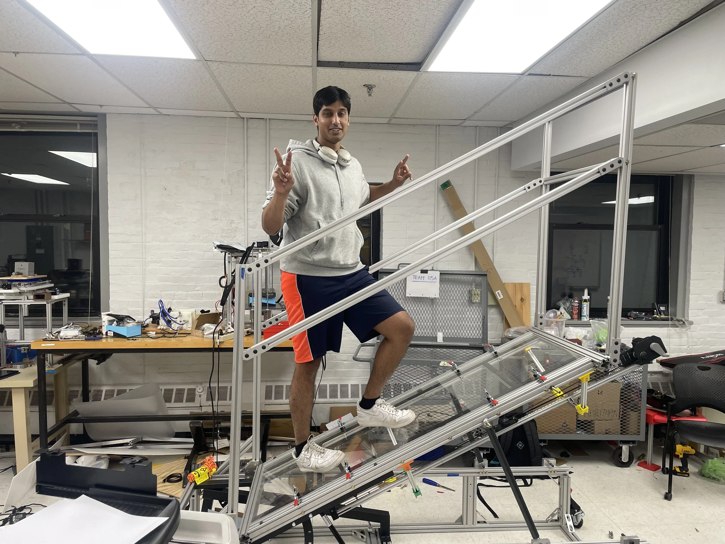 A young man in a gray hoodie and shorts standing on a ramp or staircase prototype, making peace signs with both hands, in a workshop or lab setting.