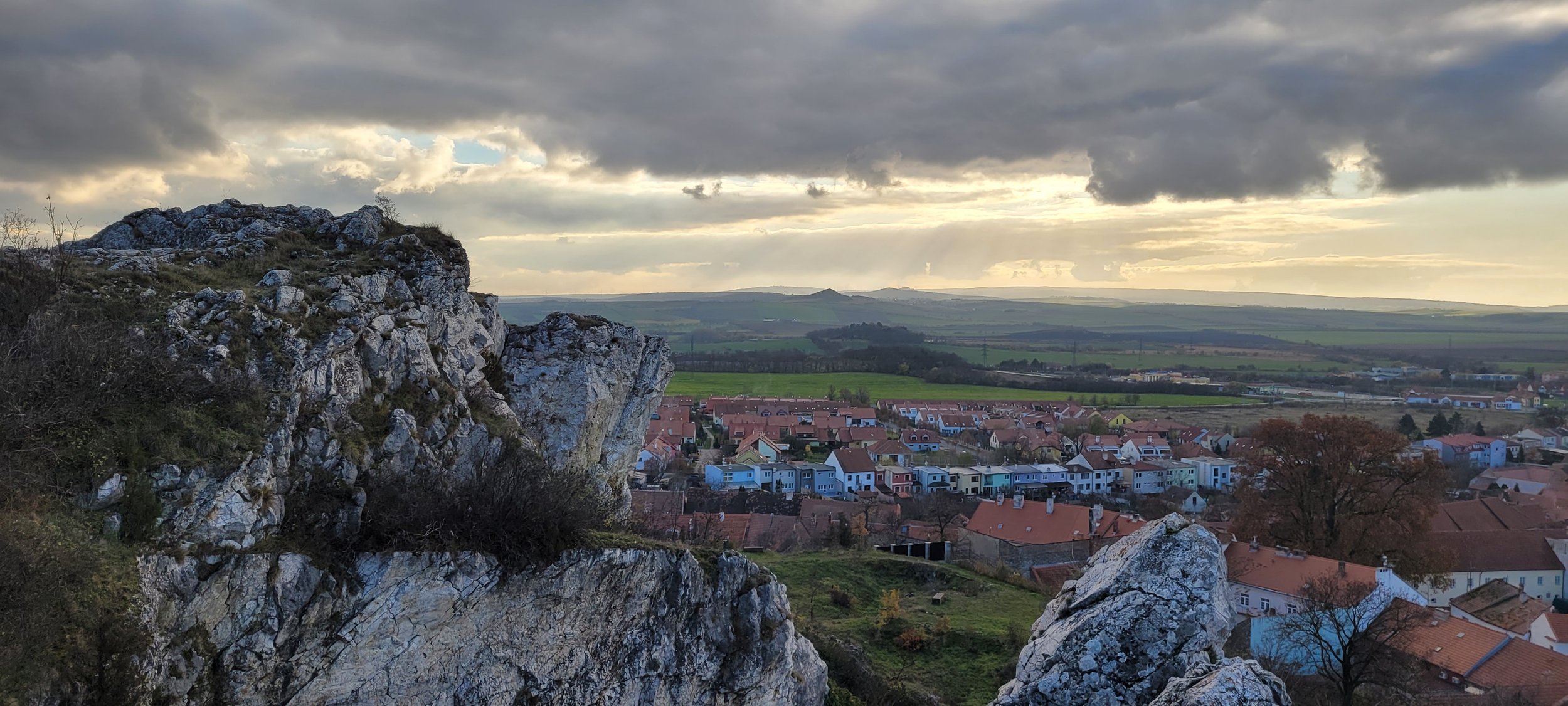 A scenic view of a town with colorful houses, large rocks in the foreground, and rolling green hills in the background under a cloudy sky.