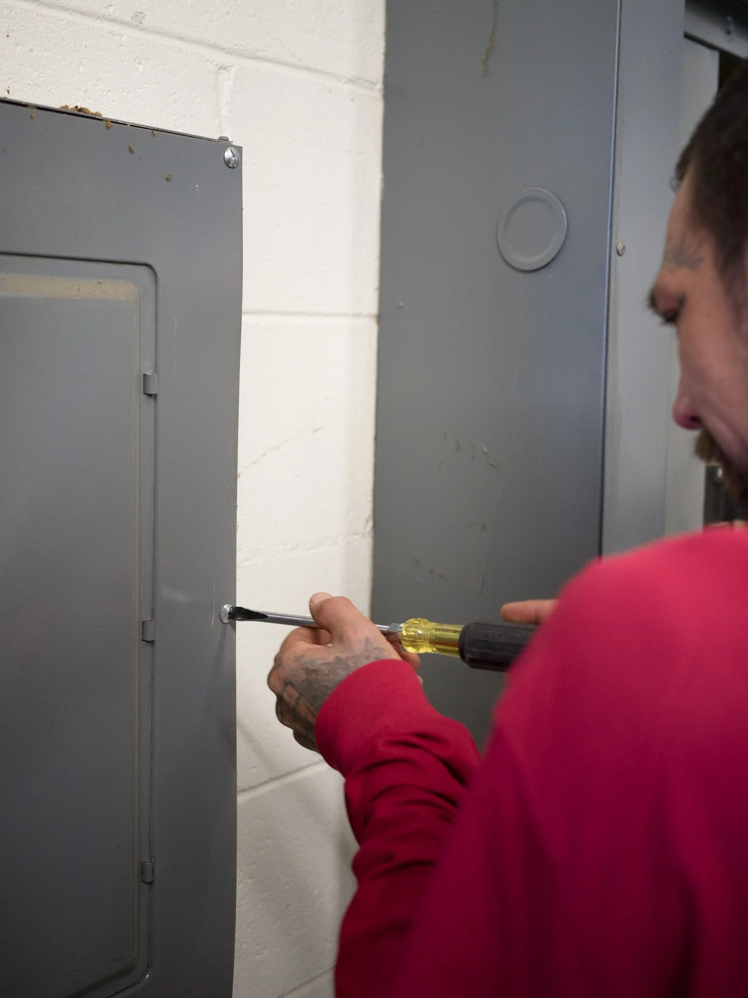 A man in a red shirt using a screwdriver to work on a gray electrical panel mounted on a white concrete wall.