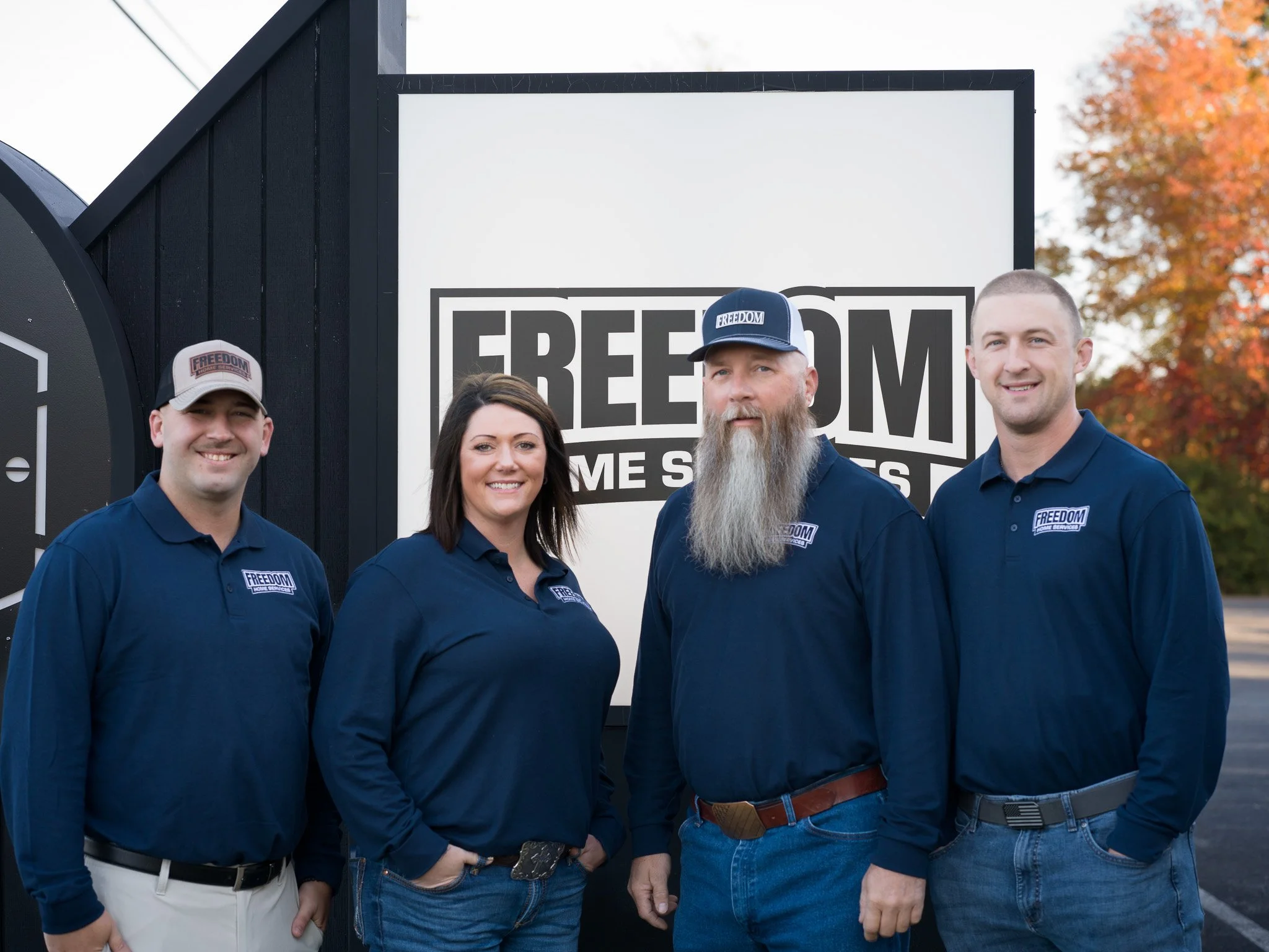 Four people standing together outdoors in front of a sign that reads 'FREEDOM' with additional text underneath. They are all wearing navy blue shirts with a logo that says 'FREEDOM' and are smiling at the camera.