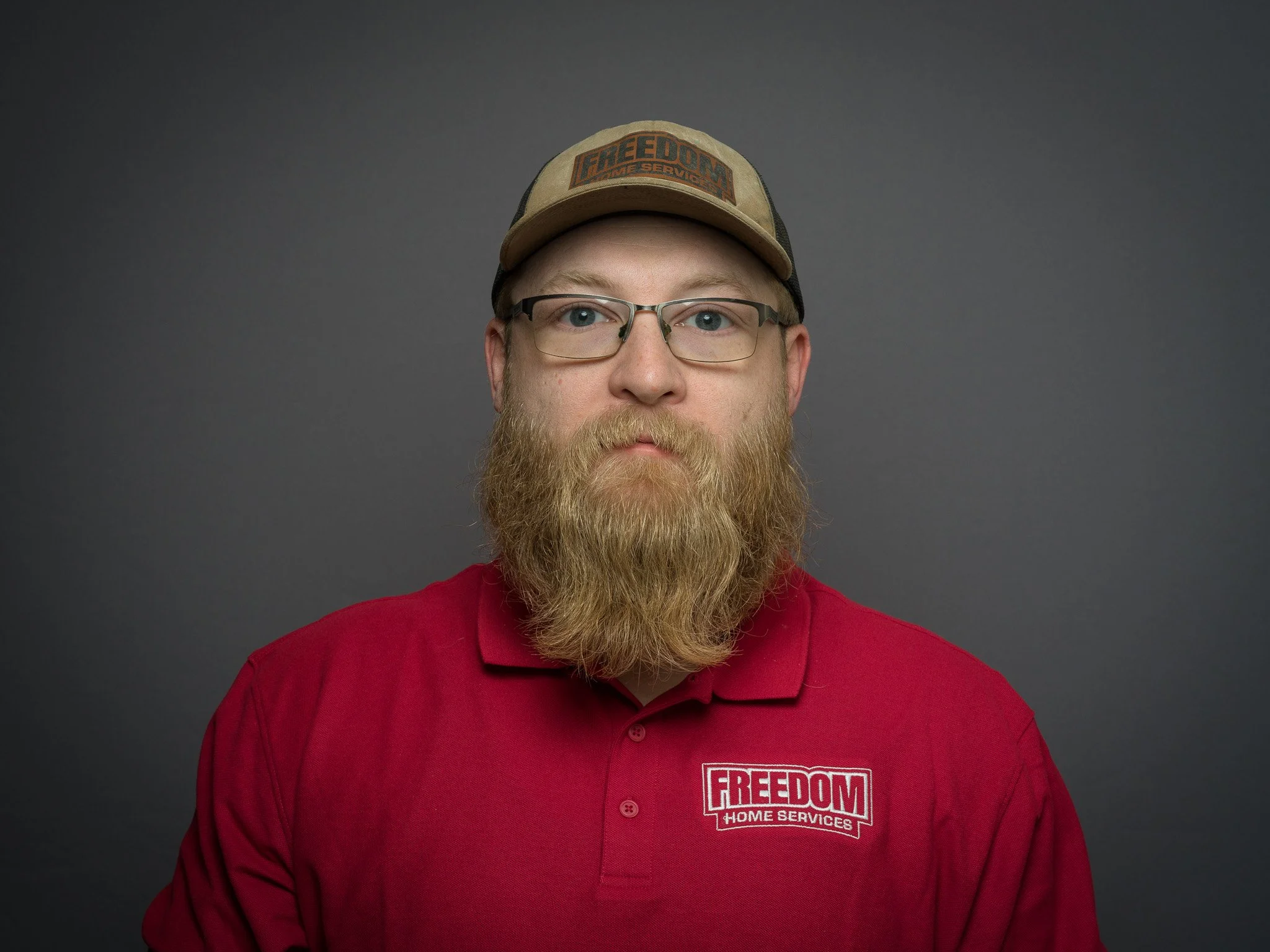 A man with glasses and a bearded face wearing a red polo shirt with a 'FREEDOM HOME SERVICES' logo and a beige cap with a 'FREEDOM HOME SERVICES' patch, posing in front of a plain dark background.