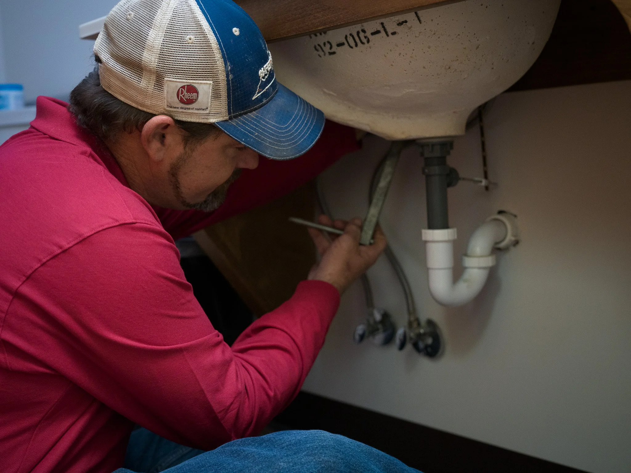 A man wearing a red shirt and a blue and beige cap working under a kitchen sink, fixing or inspecting plumbing pipes.