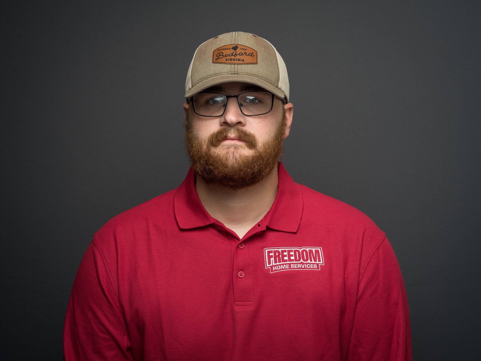 A man with glasses and a beard wearing a beige baseball cap and a red polo shirt with a "FREEDOM HOME SERVICES" logo, standing against a dark gray background.