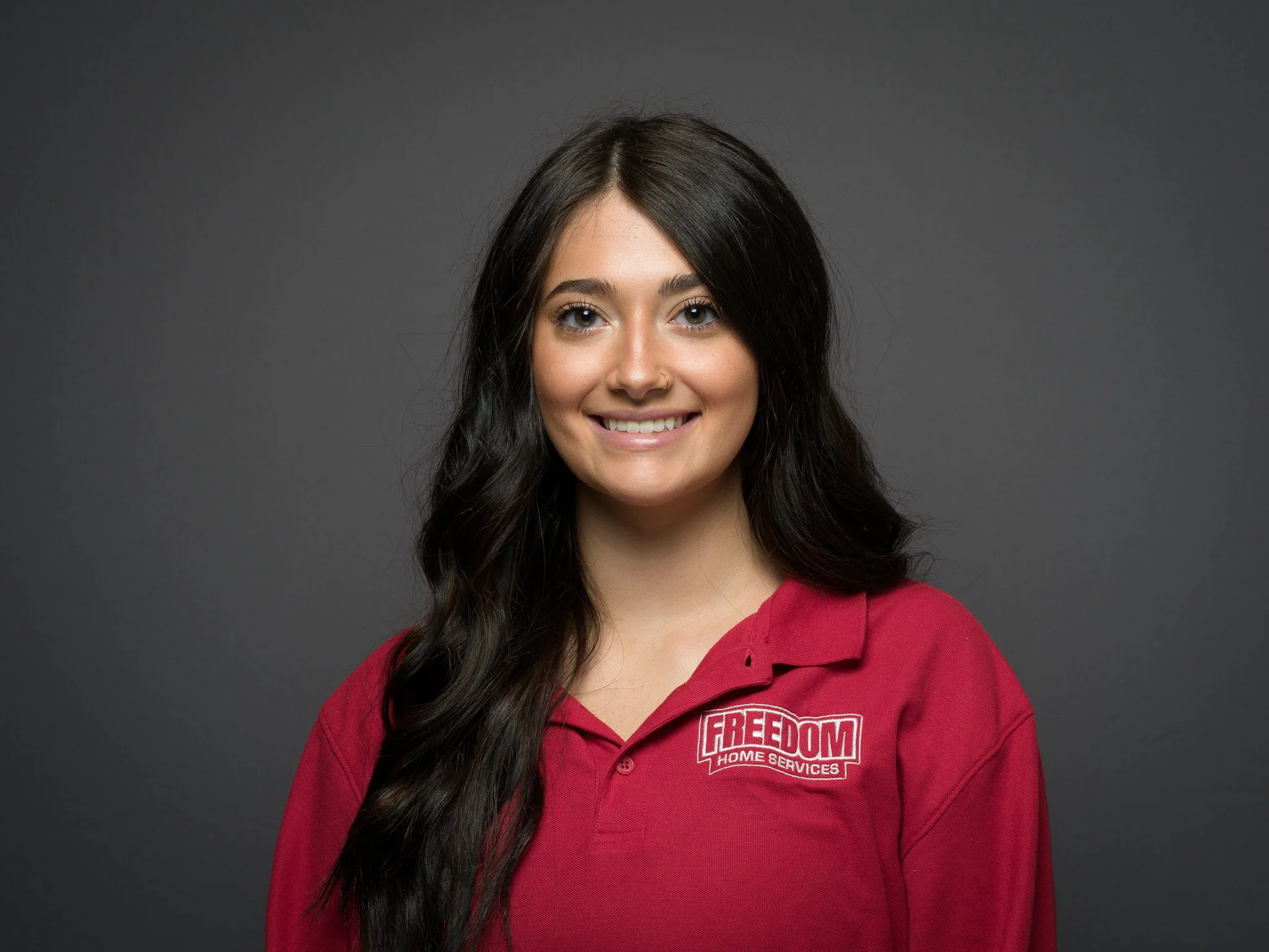 Portrait of a woman with long dark hair wearing a red polo shirt with 'FREEDOM HOME SERVICES' logo, smiling against gray background.