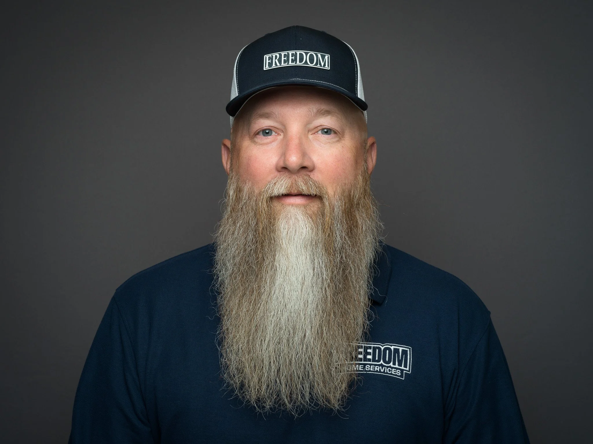 Man with long beard wearing a navy blue cap with 'FREEDOM' and a navy blue shirt with 'FREEDOM HOME SERVICES' logo, posing against a gray background.