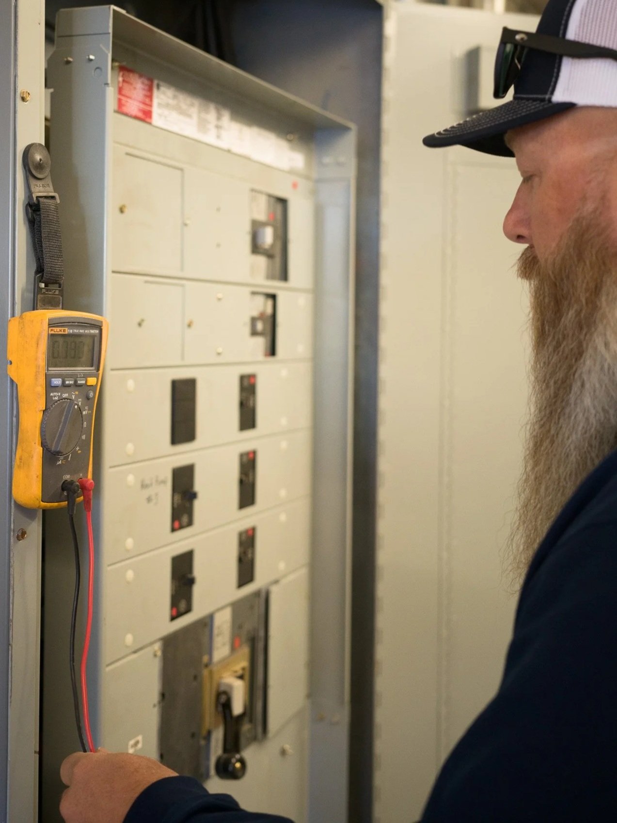 A man with a long beard and a cap using a multimeter to test electrical panels or circuit breakers.
