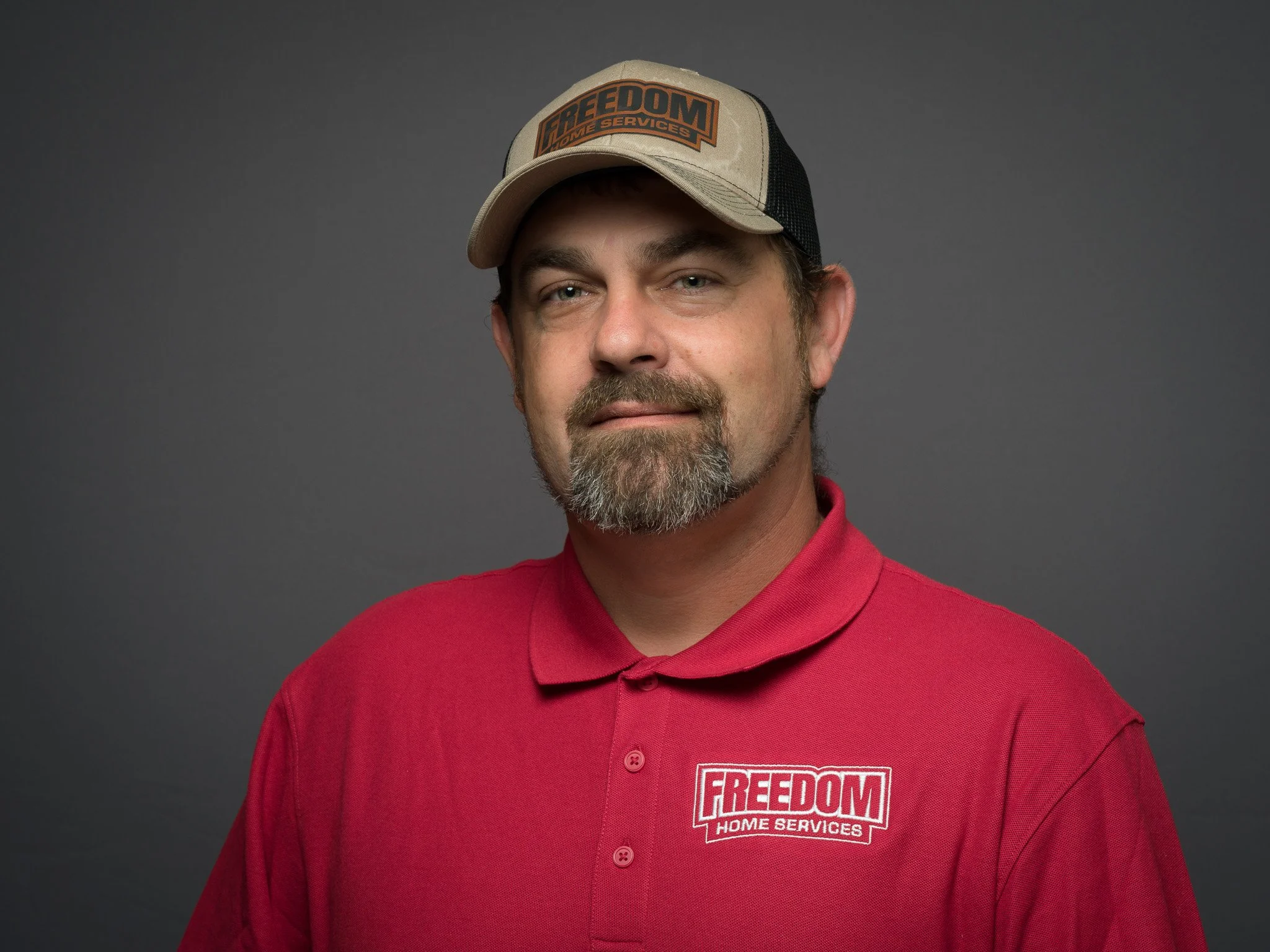 A man wearing a red polo shirt and a beige and black cap with the logo 'FREEDOM HOME SERVICES' stands against a gray background.