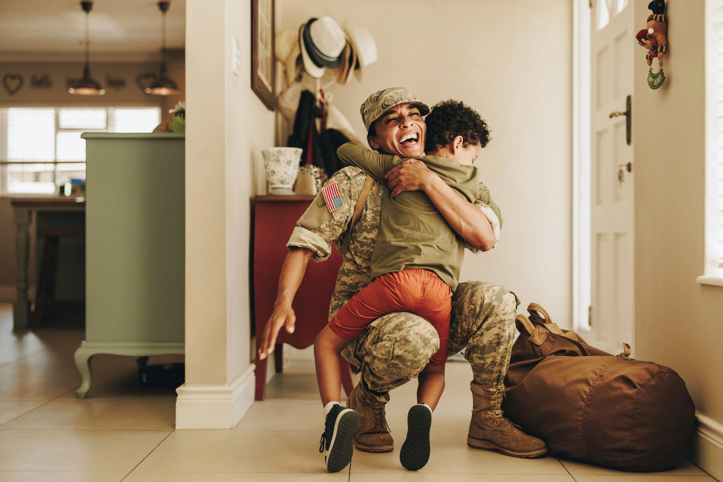 A female soldier in uniform embraces a young boy in a home entryway, both smiling joyfully.