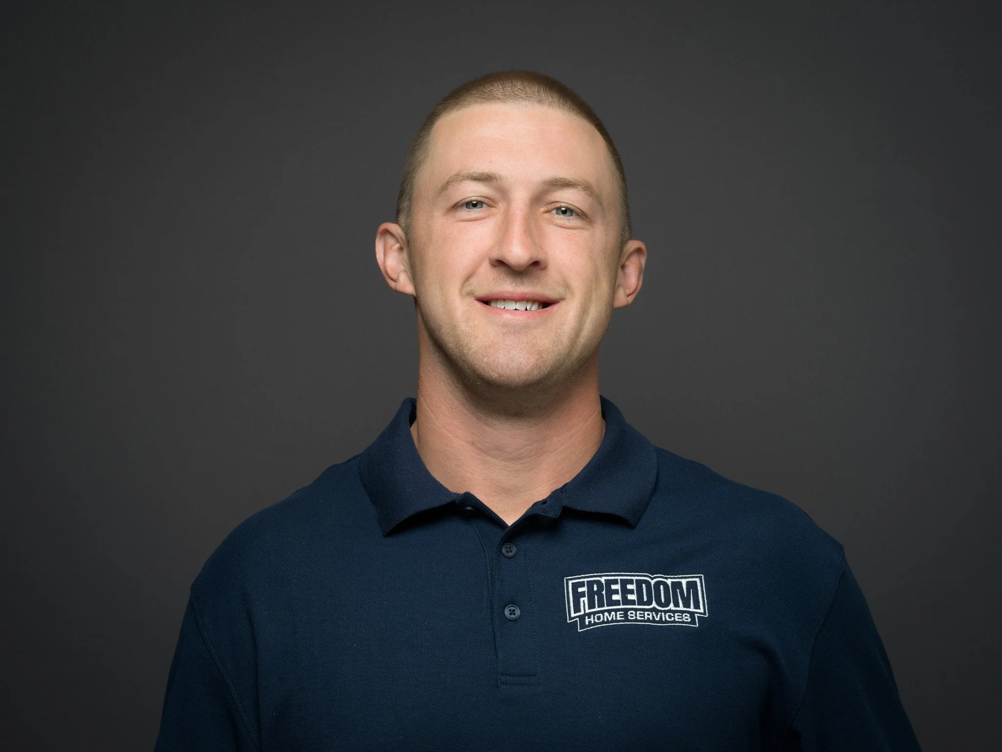 A smiling man in a navy polo shirt with a logo that reads 'FREEDOM HOME SERVICES' on the left chest area, against a dark background.