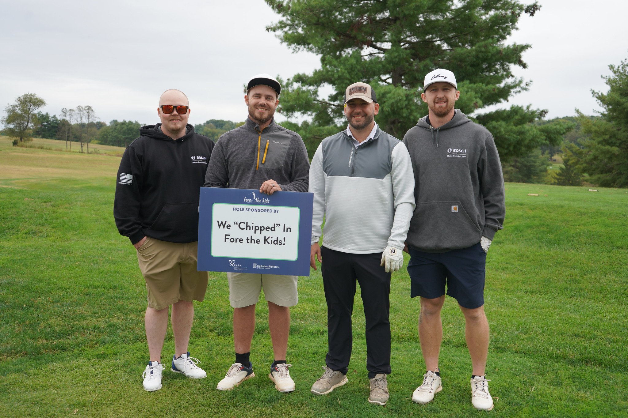 Four men standing on a golf course holding a sign that reads 'We 'Chipped' In Fore the Kids!' with trees and grass in the background.