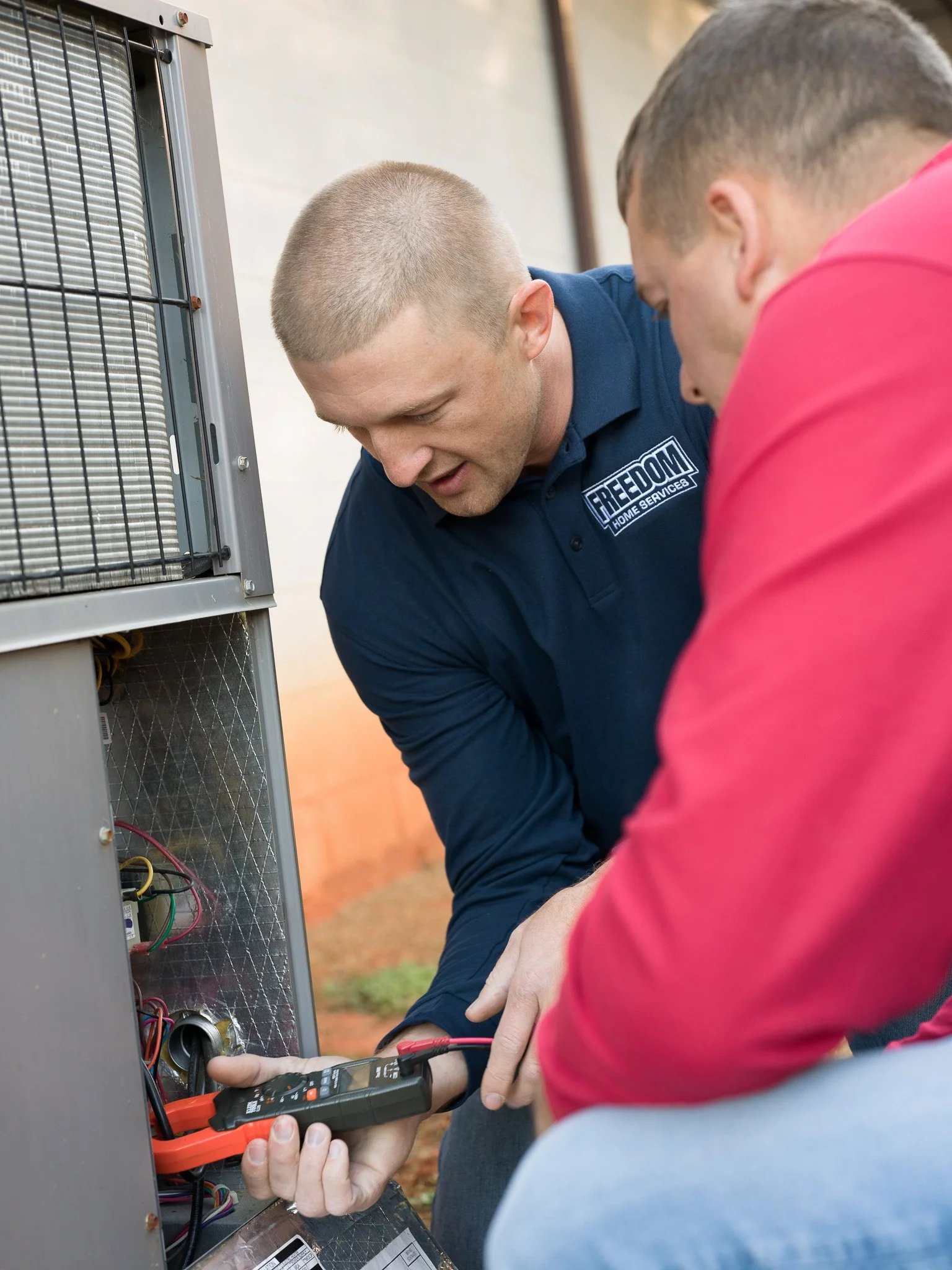 Two technicians perform maintenance on an outdoor HVAC unit, one using a multimeter to check electrical components, another observing.