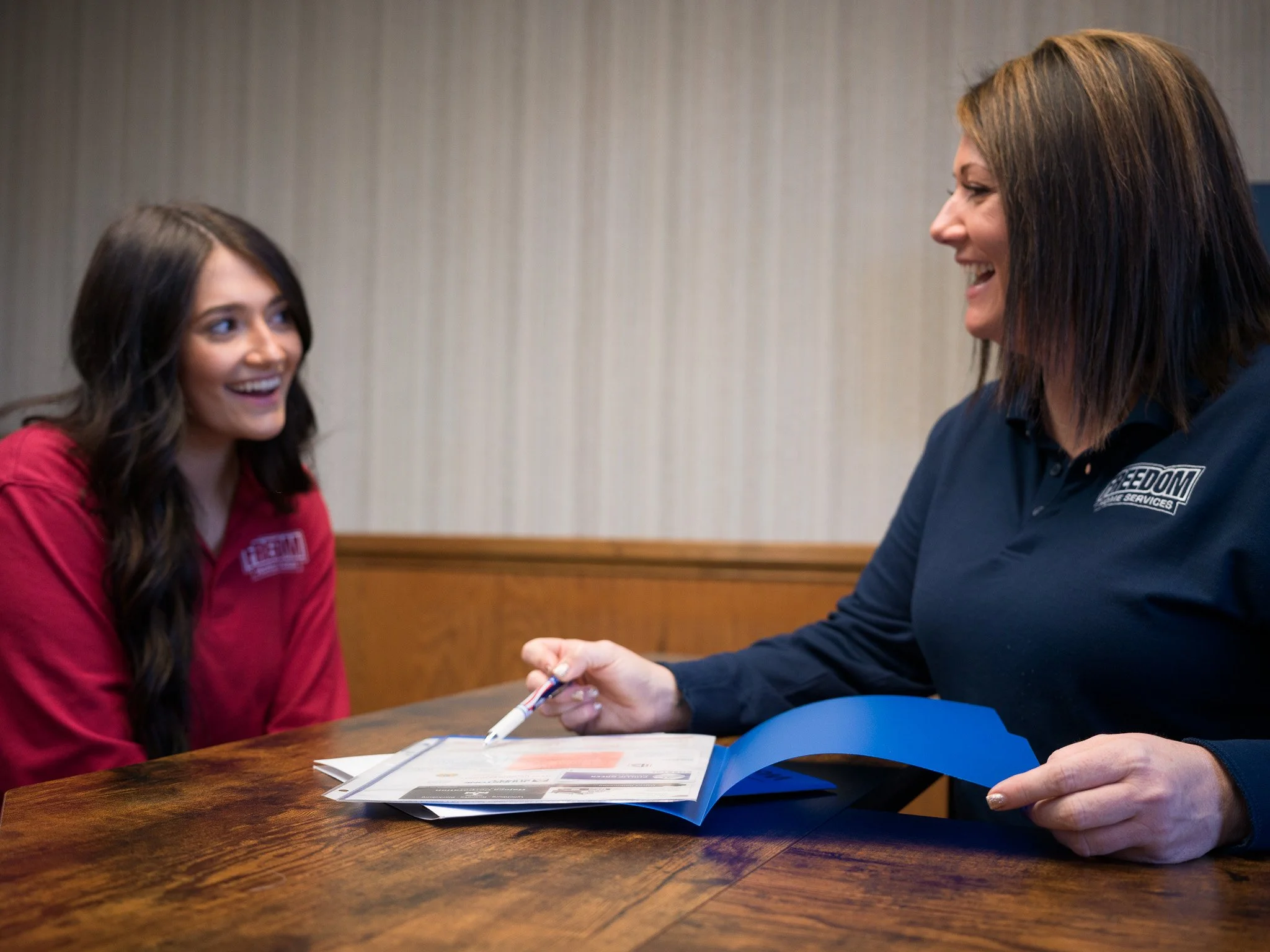 Two women sitting at a wooden table, smiling and talking. One woman is wearing a red shirt, the other a navy blue shirt with a company logo. The woman in blue is holding a pen and pointing at a pamphlet or brochure.