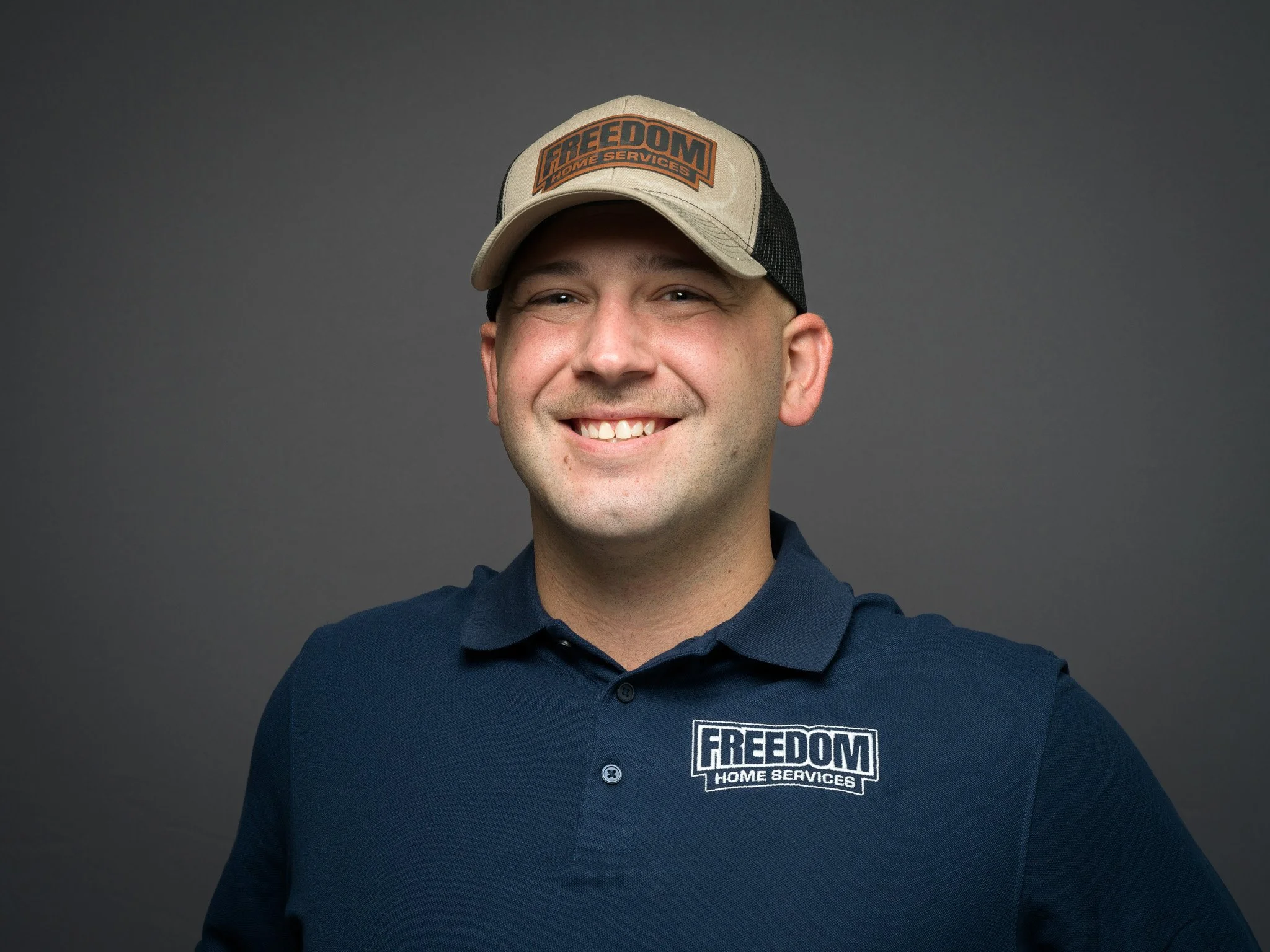 Smiling man wearing a navy polo shirt and a beige trucker cap with 'FREEDOM HOME SERVICES' logo, against a gray background.