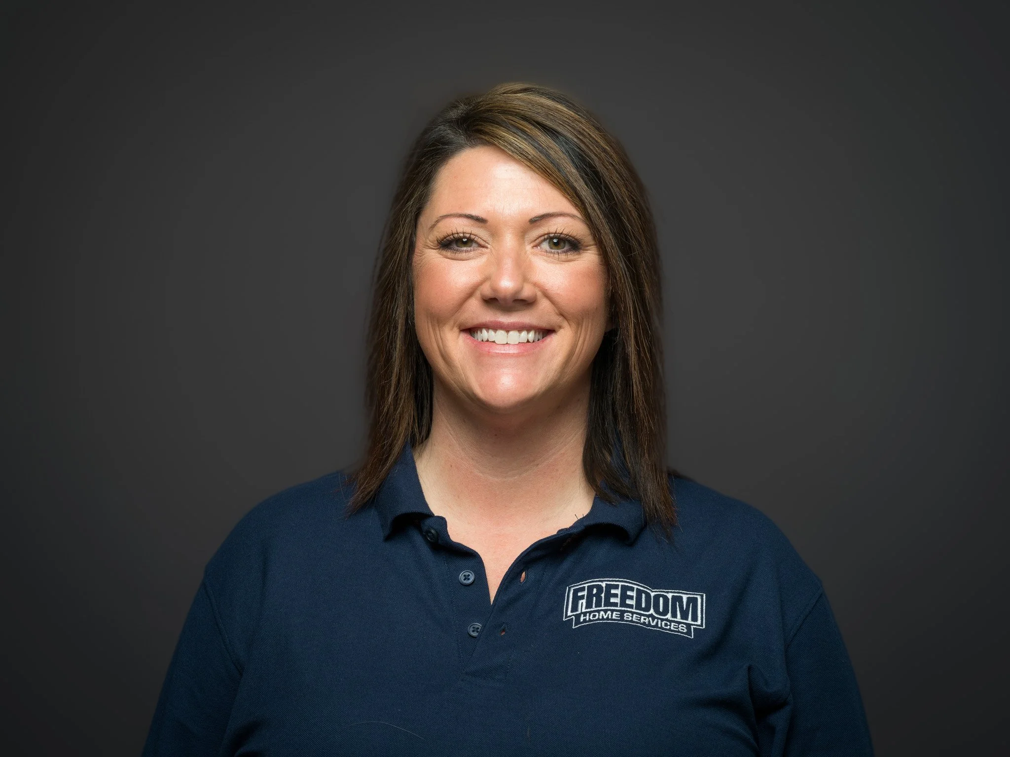 A smiling woman with shoulder-length brown hair, wearing a navy blue polo shirt with a logo that reads 'FREEDOM HOME SERVICES,' against a dark gray background.