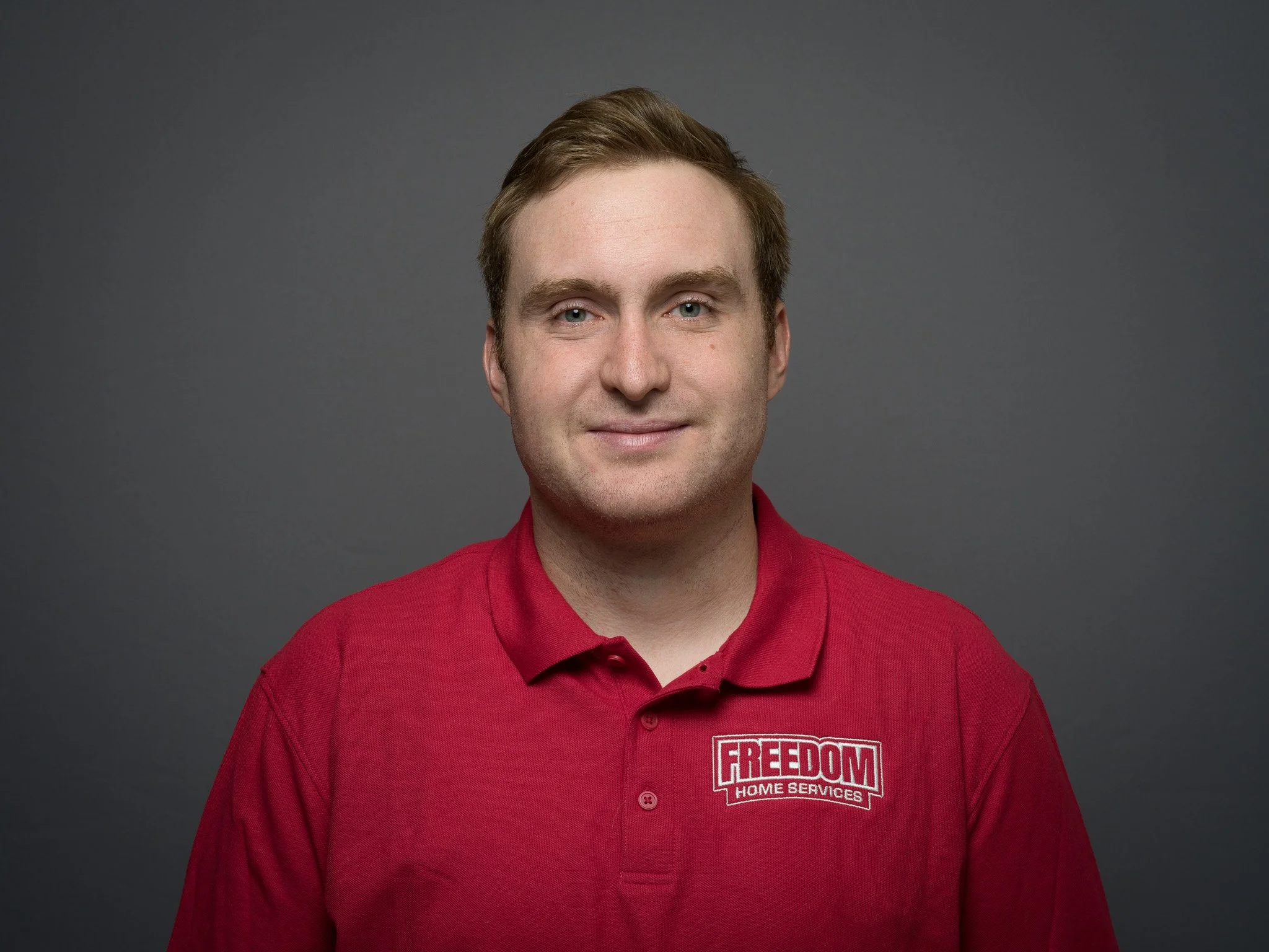 A man with light skin, brown hair, and blue eyes wearing a red polo shirt with 'FREEDOM HOME SERVICES' logo embroidered on it, standing against a plain gray background.