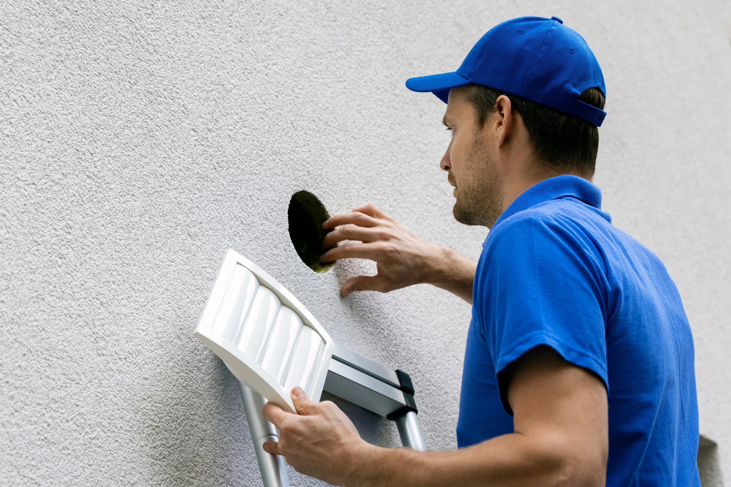 A man in a blue uniform and cap repairing a textured white wall, using a scaffold and holding a black patch.