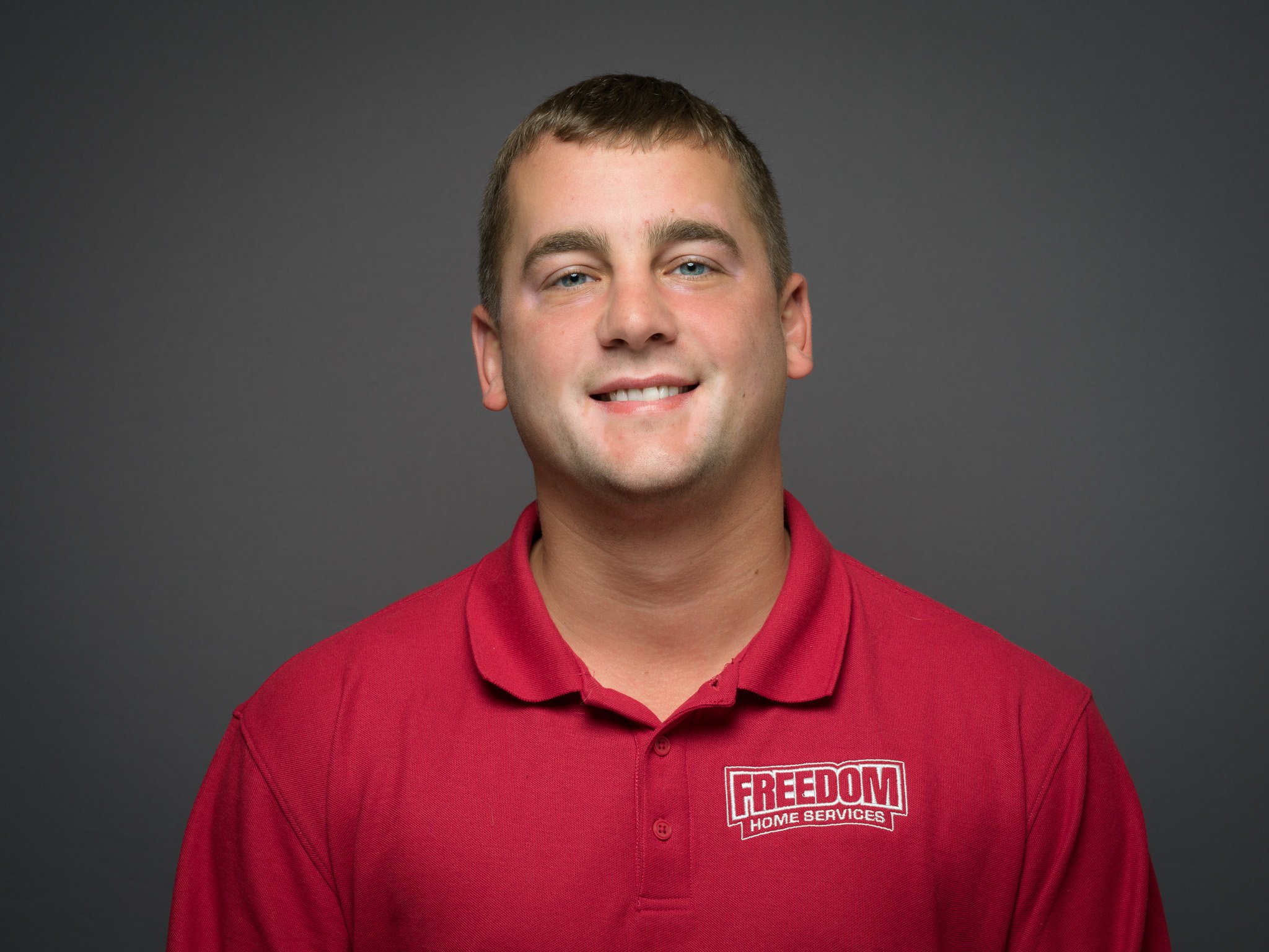 A young man with short brown hair and blue eyes, smiling, wearing a red polo shirt with the logo "FREEDOM HOME SERVICES" on it, against a dark gray background.
