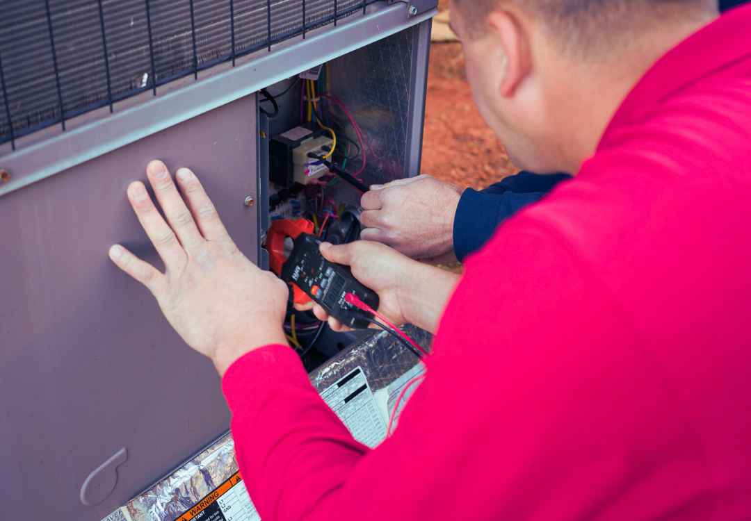 A technician wearing a red jacket using a digital multimeter to test electrical components inside an HVAC unit outdoors.