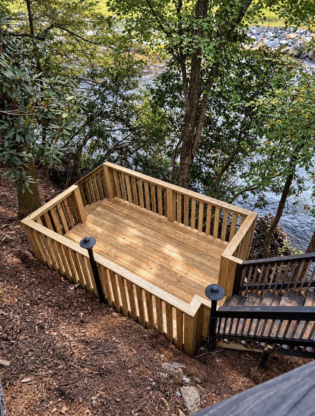 Wooden deck with outdoor lighting, overlooking a river, surrounded by trees and natural landscape.